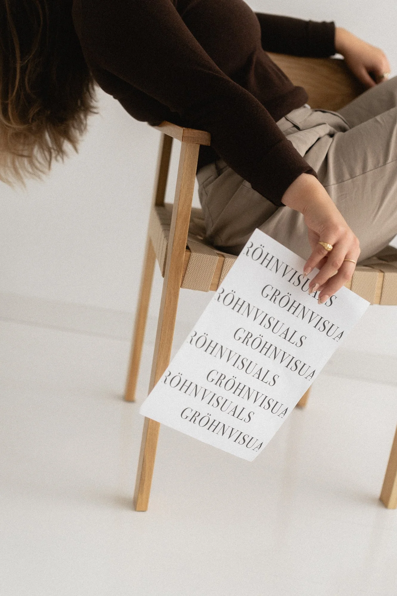Henkilö istuu puisella tuolilla ja pitää paperia, jossa on tekstiä 'RÖHNWISUALS GRÖHNVISUA RÖHNVISUALS GRÖHNVISUA RÖHNVISUALS GRÖHNVISUA RÖHNVISUALS'.