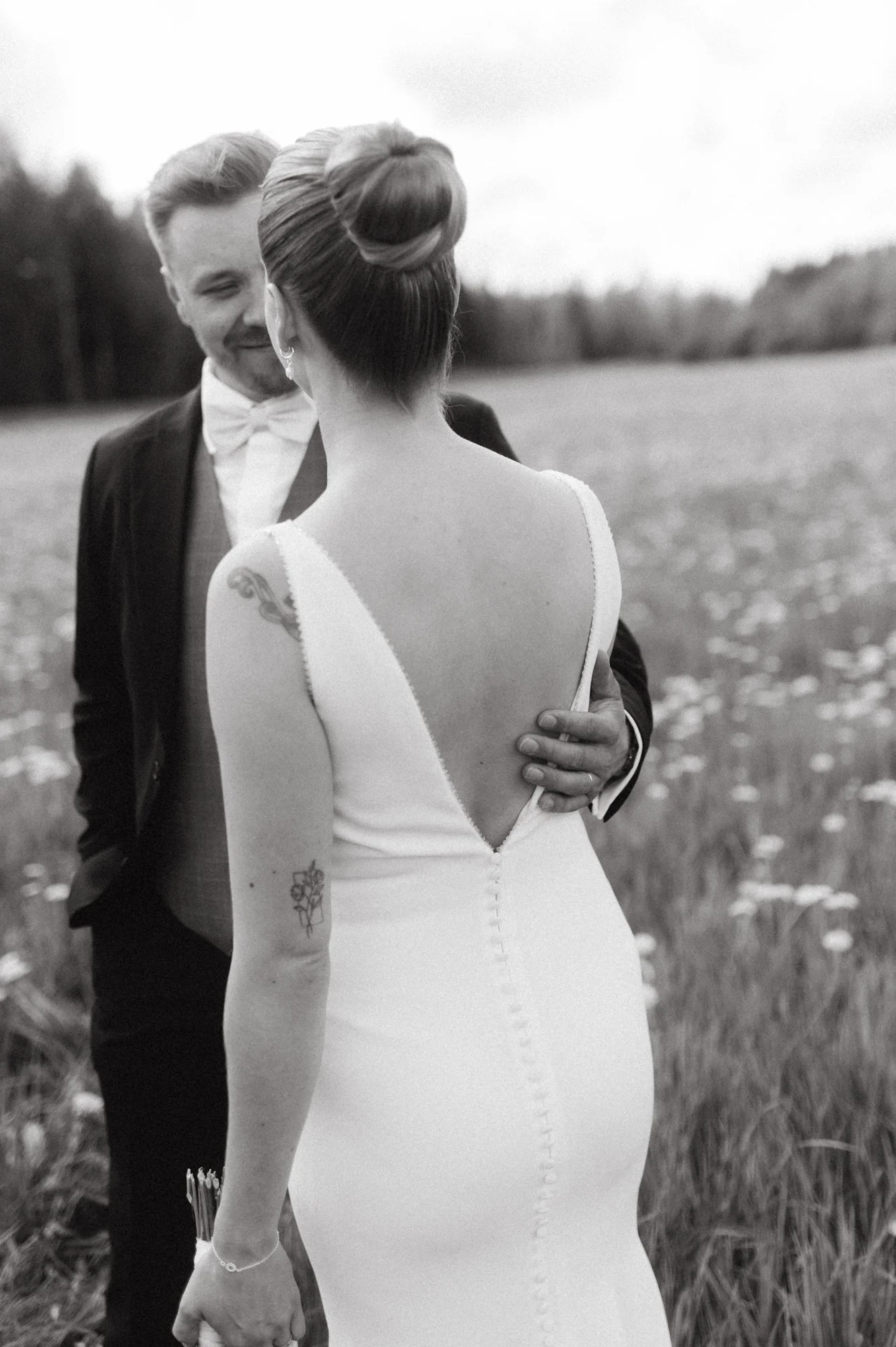 Morsiamen ja sulhanen hymyilevät toisilleen ulkona, morsian pukeutunut valkoiseen hääpukuun, sulhanen pikkutakkiin ja puseroon, taustalla ruohoa ja metsää, mustavalko