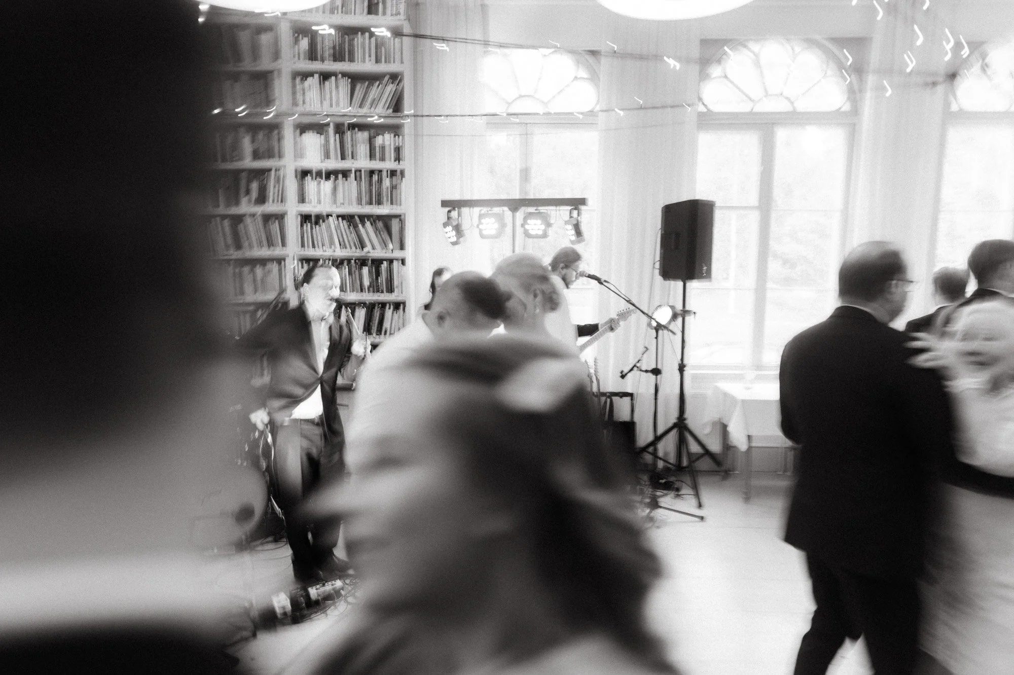 Mustavalkoinen kuva ihmisistä tanssimassa ja musiikkia soittavasta bändistä, salin taustalla kirjahylly ja suuret ikkunat.