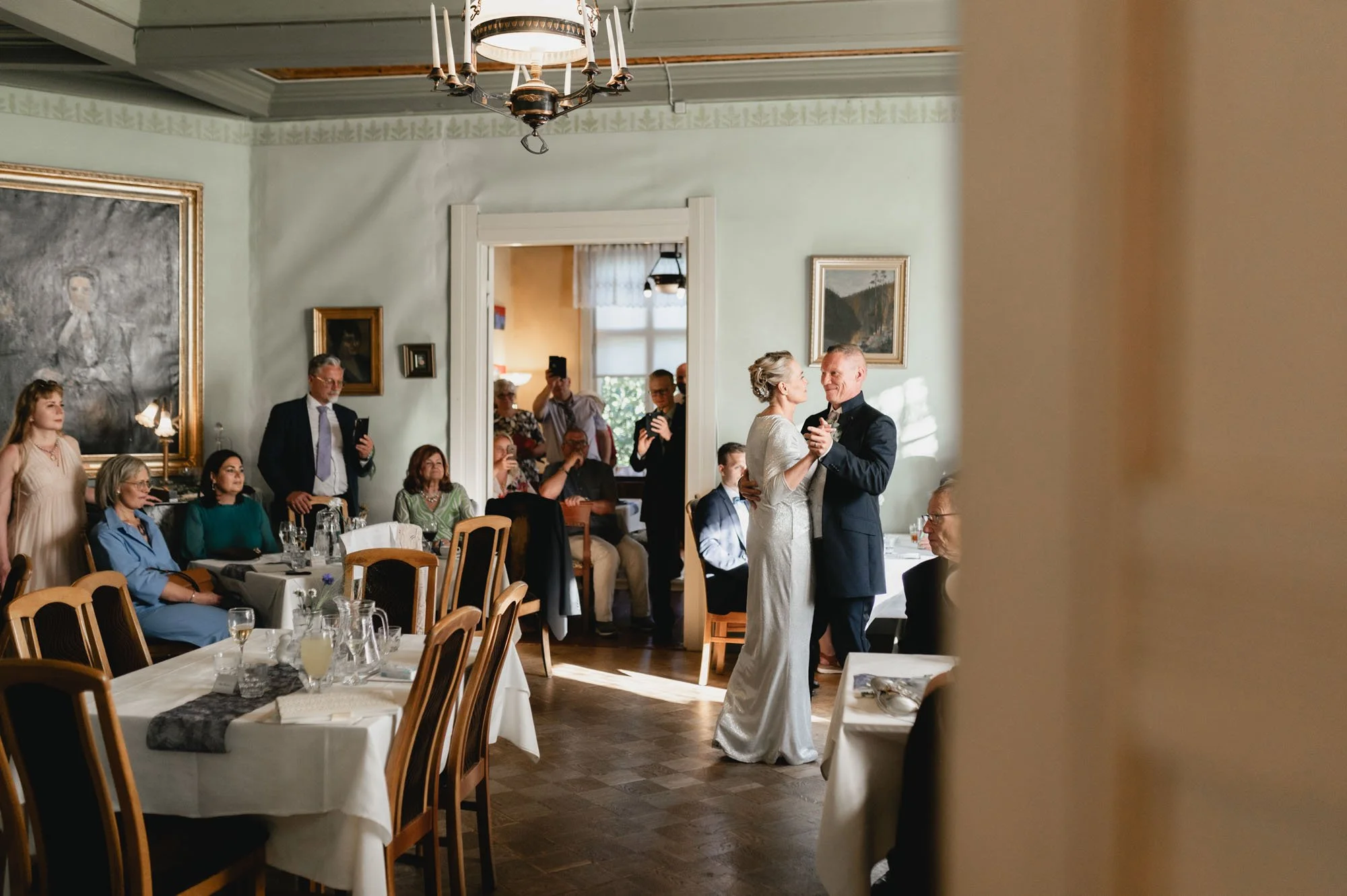Hääpari tanssimassa ruokasalin keskellä, vieraat istumassa ja seuraamassa, taulut seinällä ja kattokruunu katossa