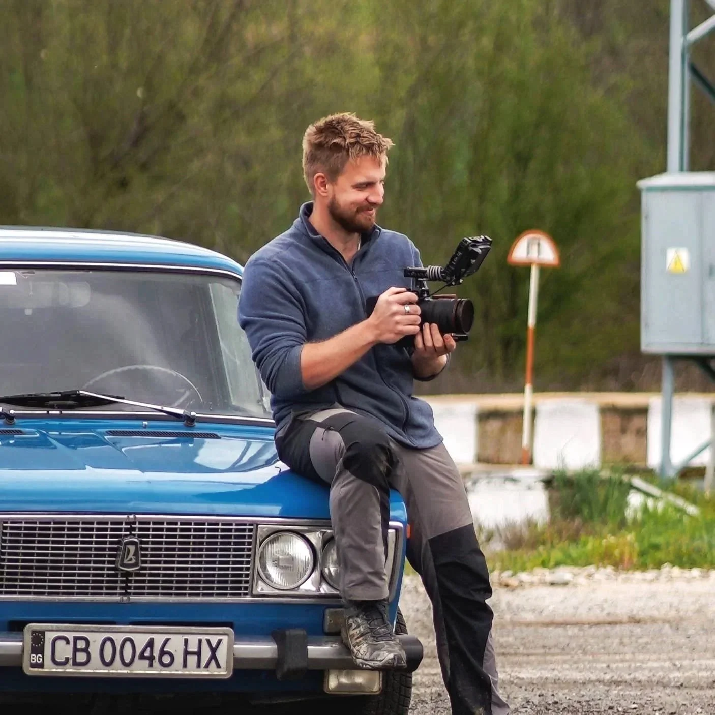 A man with a beard and short hair, dressed in a blue jacket and gray outdoor pants, is sitting on the hood of a vintage blue car with a Bulgarian license plate, holding a professional camera with an external microphone, outdoors in a rural setting.
