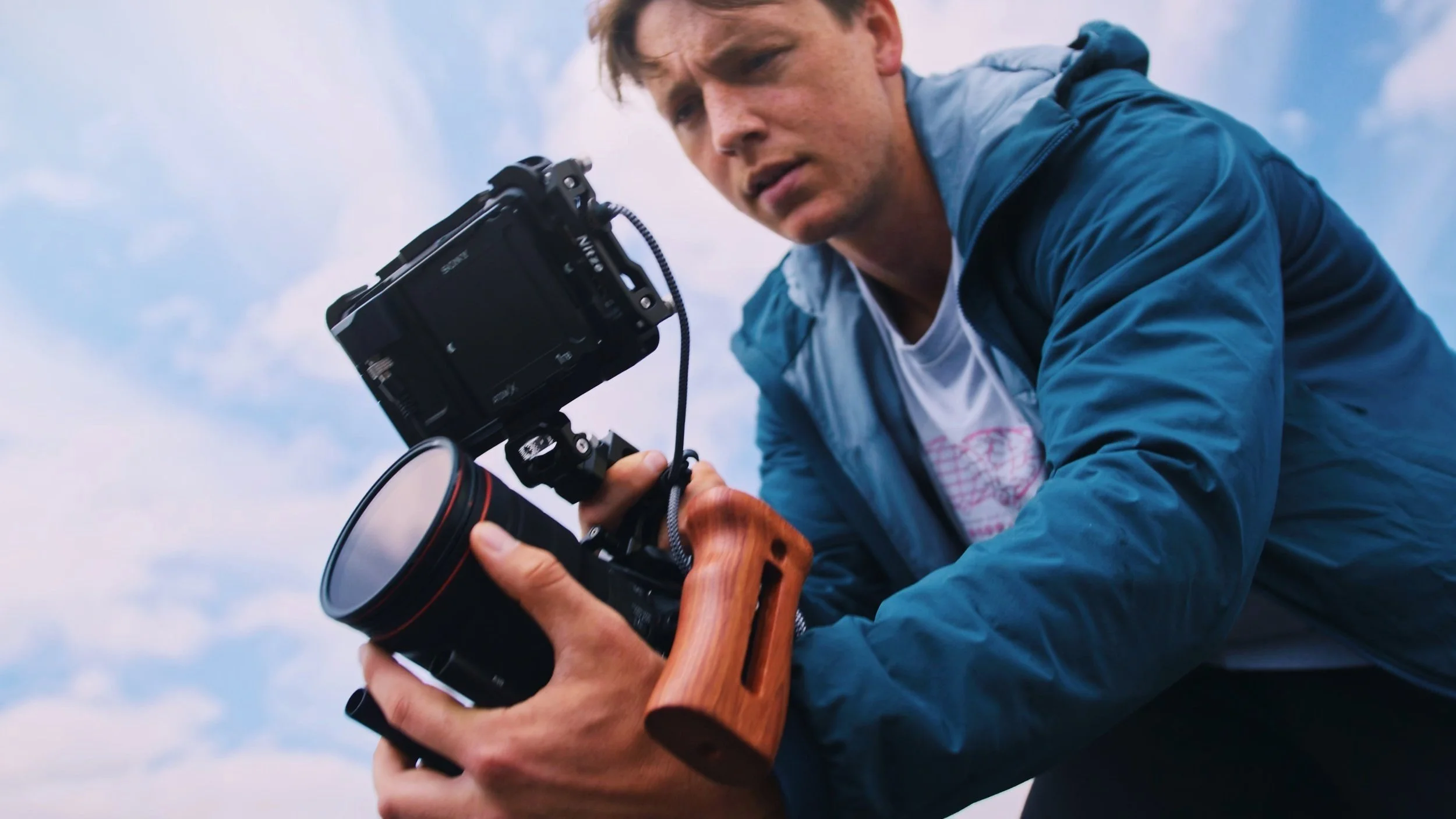 A man in a blue jacket is holding a professional camera with a wooden handle, aiming it outdoors against a cloudy sky.