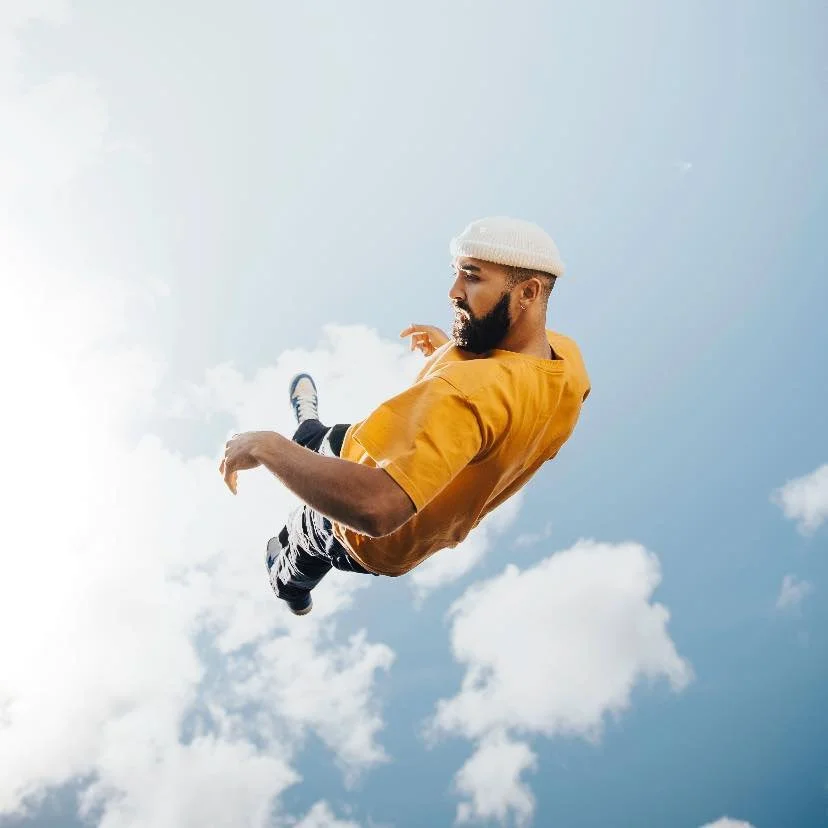 A man wearing a yellow shirt, white beanie, and sneakers appears to float or fall against a cloudy blue sky.
