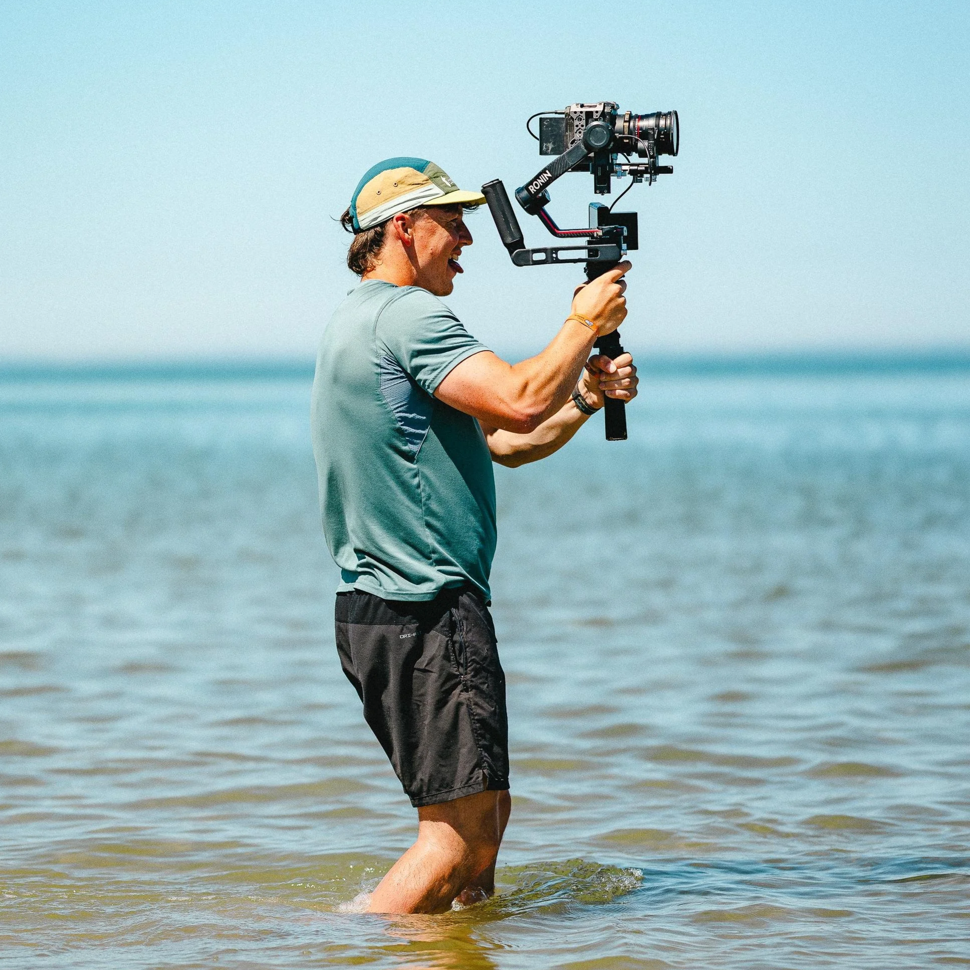 Man filming with a camera stabilizer in shallow water at the beach.