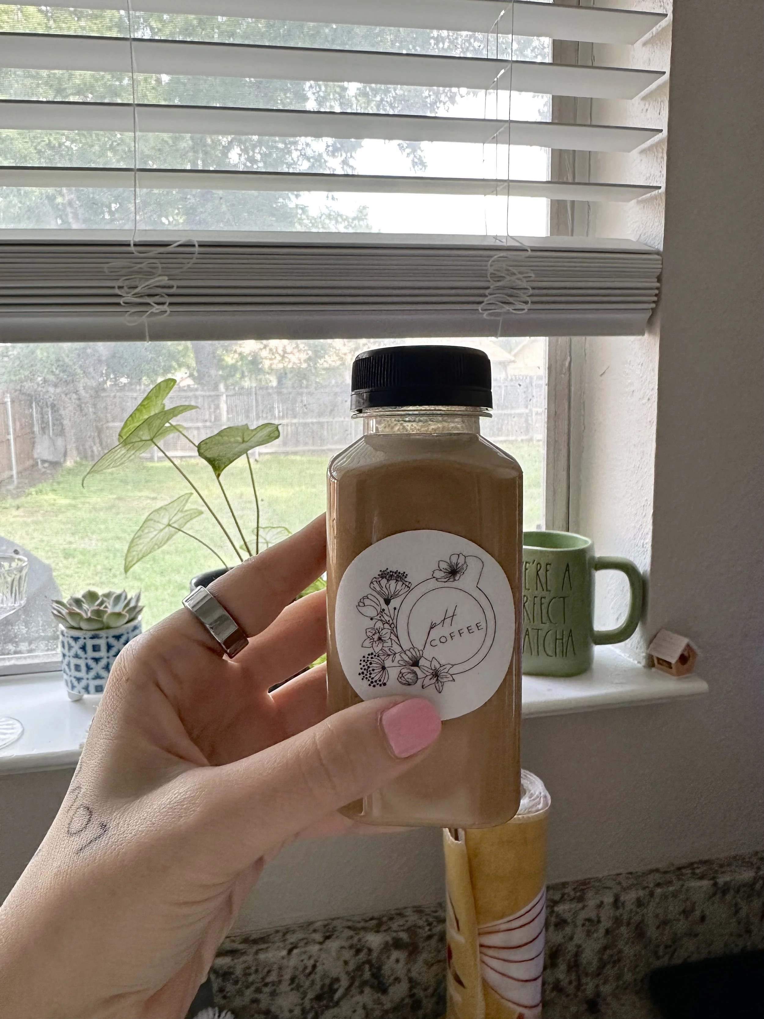 A person holding a bottle of peach coffee flavoring in front of a window with blinds, with a plant, a green mug, and a small decorative house on the windowsill.