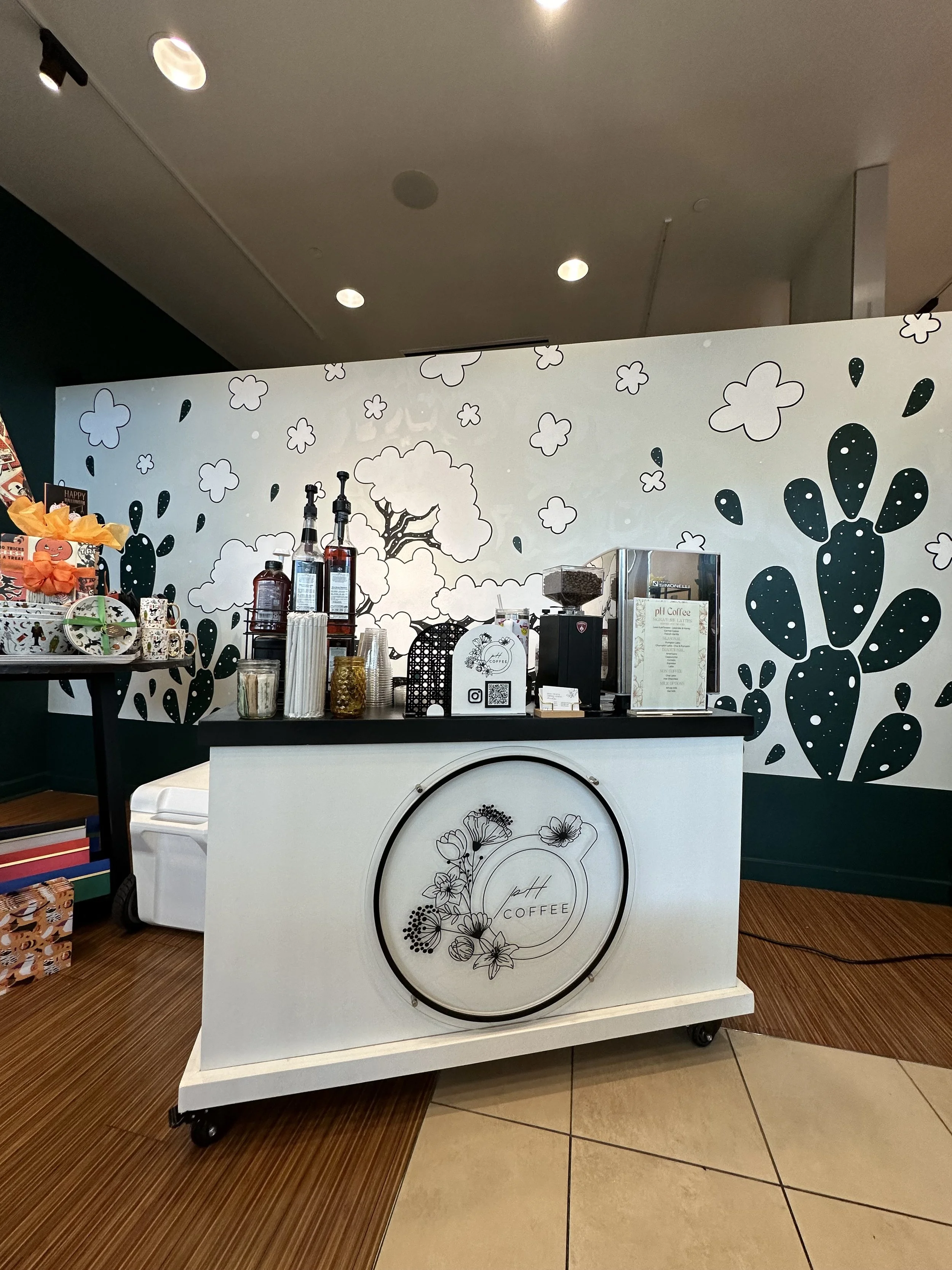 A white coffee cart with floral design, set up in a shopping mall, with coffee syrups, cups, and a coffee machine on top. The background features a black and white wall with cloud and cactus illustrations.