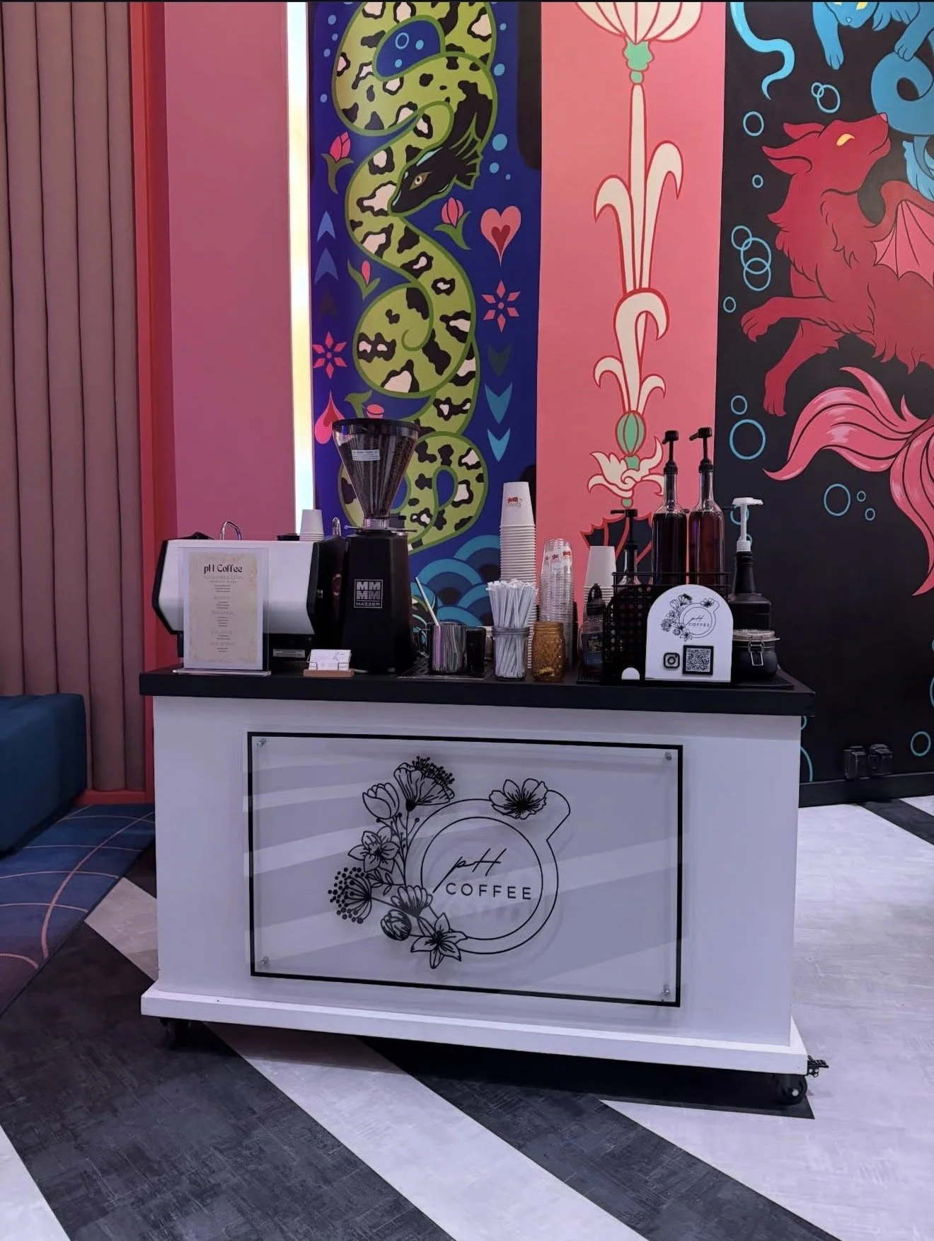 A coffee stand with a white front panel featuring a black line drawing of flowers and the words 'PH Coffee'. Behind the stand, there is a colorful, artistic wall with abstract designs, including a snake, a dragon, and decorative patterns in bright colors. The counter has a coffee grinder, cups, bottles, and other supplies.