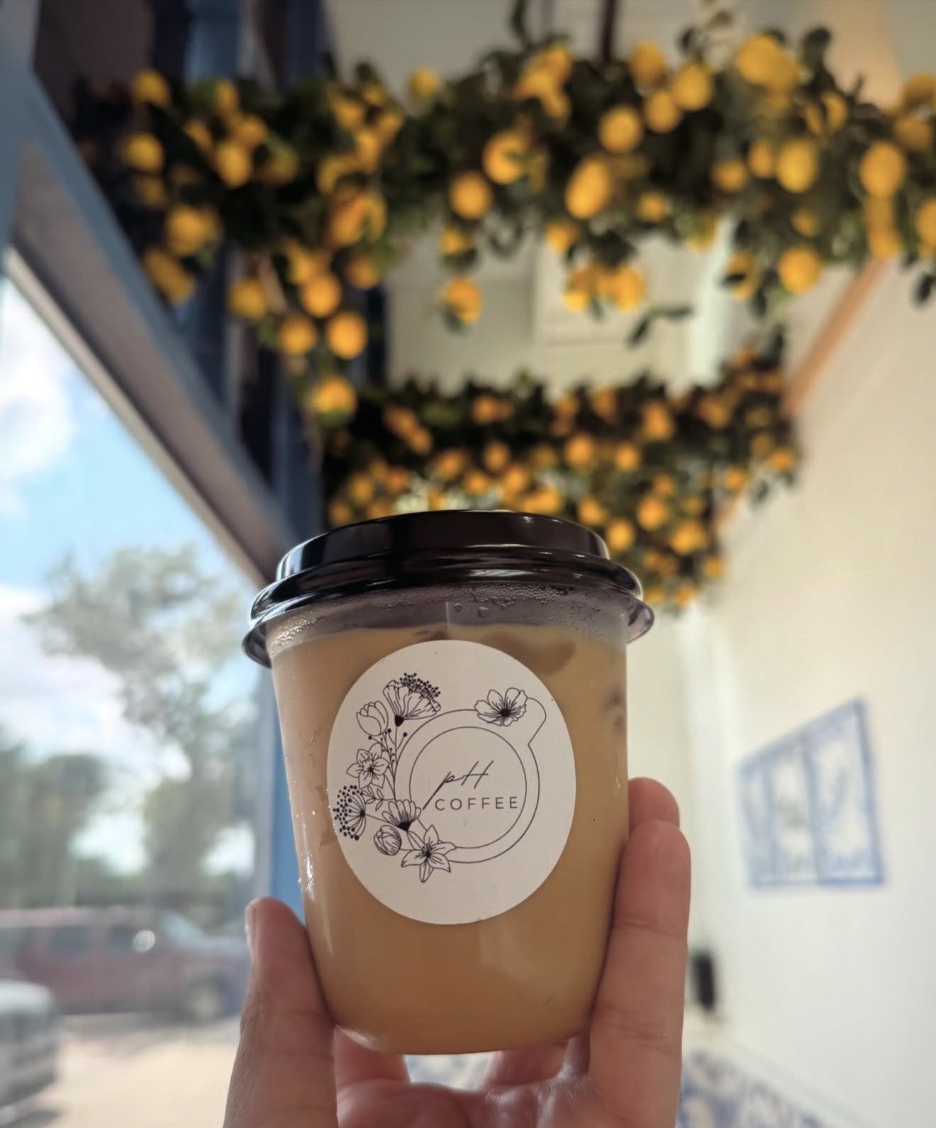 A hand holding a plastic cup of coffee with a black lid, labeled 'pnt COFFEE' with floral design, in front of a window with a view of trees and a building, and a blurred background of yellow flowering bushes outside.