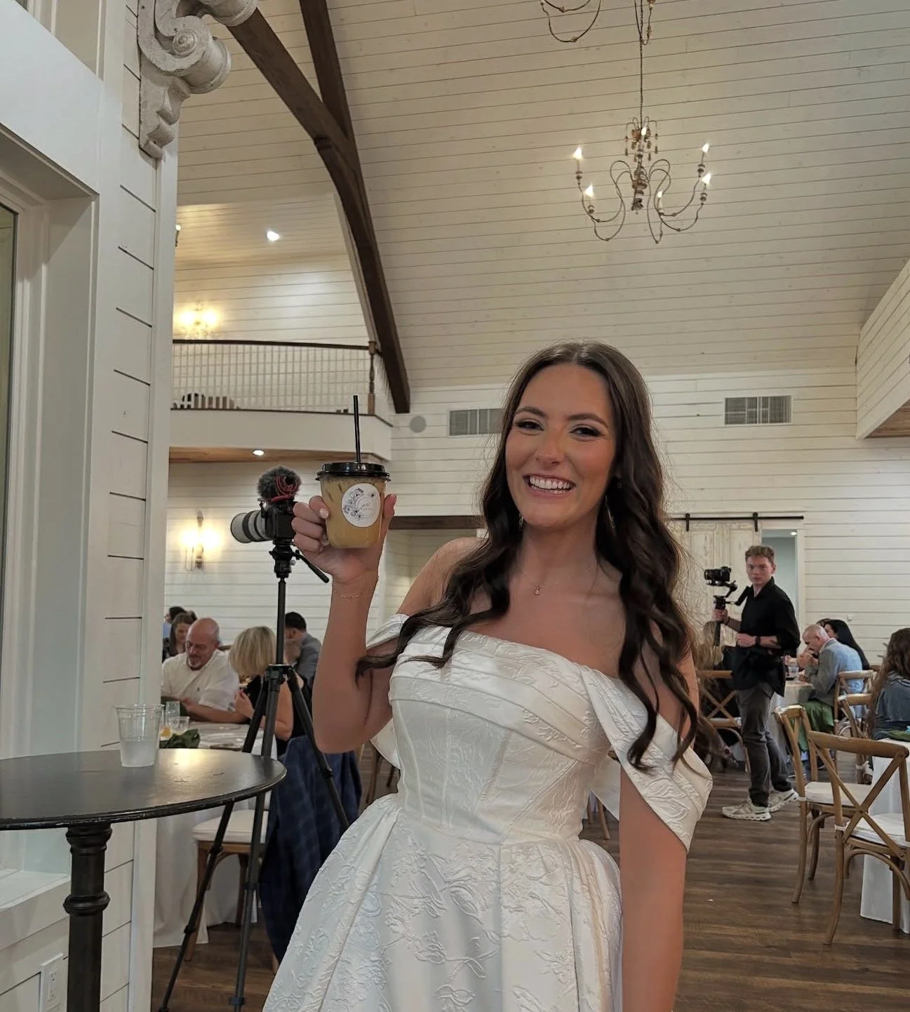 A woman in a white dress smiling and holding a to-go coffee cup with a straw, inside a decorated event space with chandelier lighting and people seated at tables.