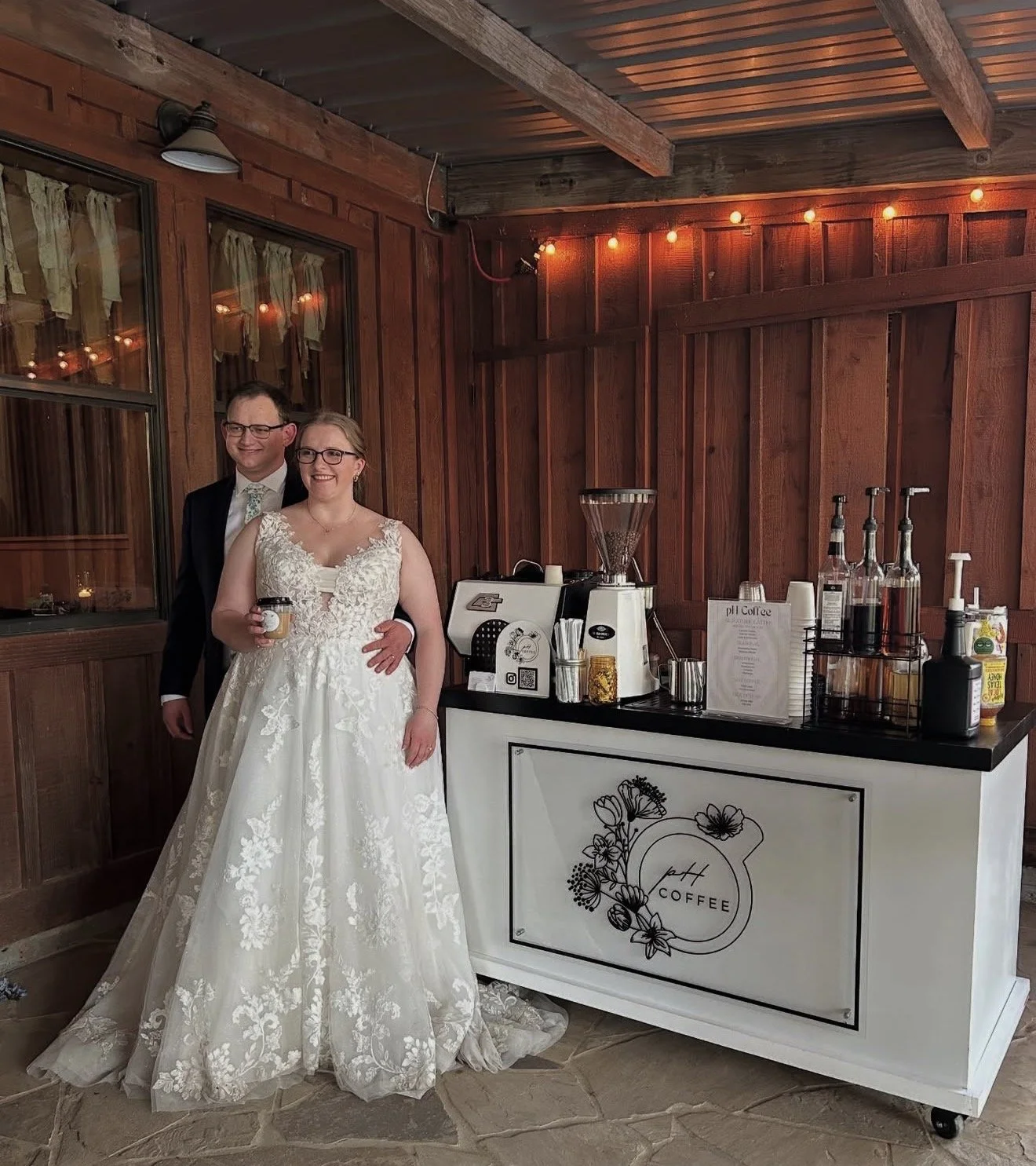 A bride and groom standing by a coffee station in a rustic indoor venue with wooden walls and string lights. The bride is wearing a white lace wedding dress, holding a drink, and the groom is in a dark suit with a light-colored tie.