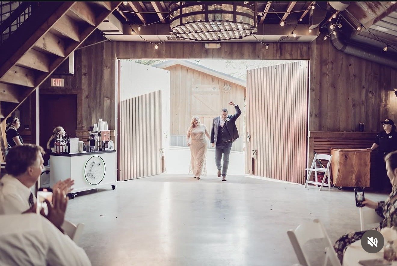 A couple walks in and out of a barn at a wedding ceremony, with guests watching and taking photos. The barn has wooden walls and open doors, with string lights hanging from the ceiling.