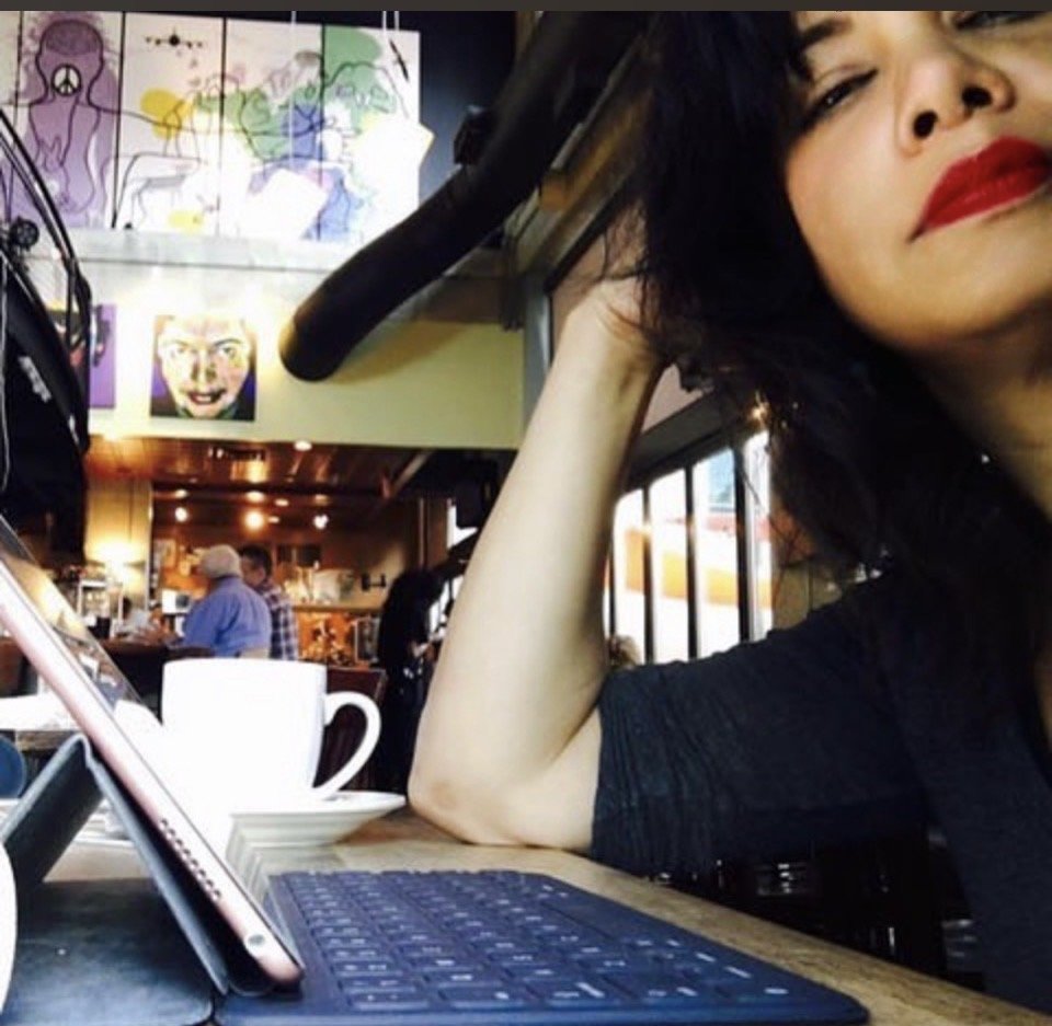 Young woman with dark hair and red lipstick sitting in a cafe, resting her head on her hand, with a tablet and coffee cup on the table in front of her. There are colorful artwork and other people in the background.