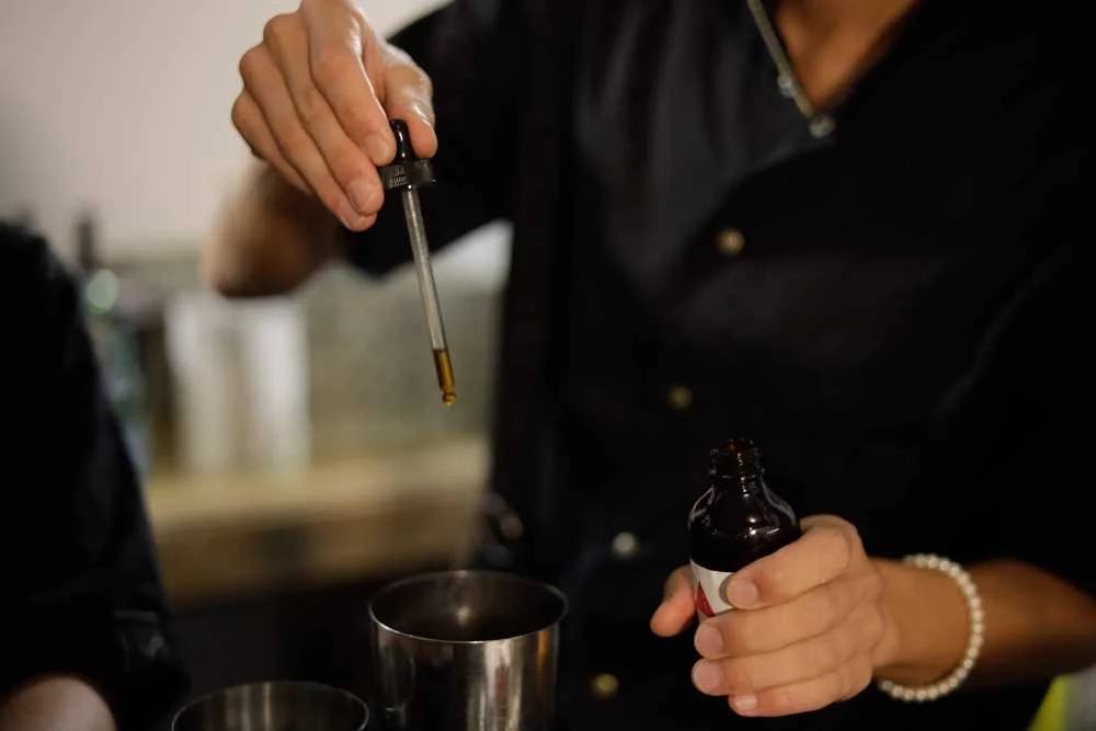 Person holding a dropper filled with a liquid, with a small brown bottle and metal cups nearby.