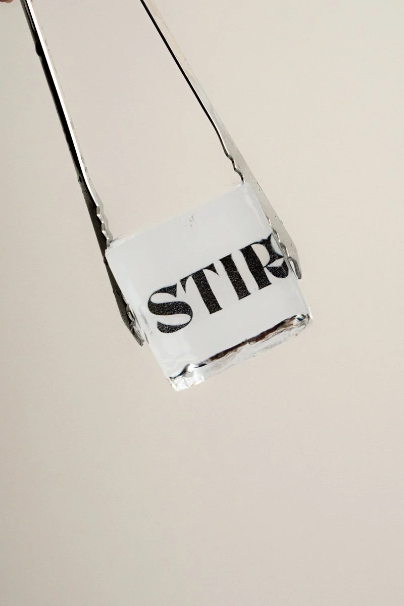 Close-up of a glass piece with the word 'STIR' printed on it, hanging downward against a plain, light-colored background.