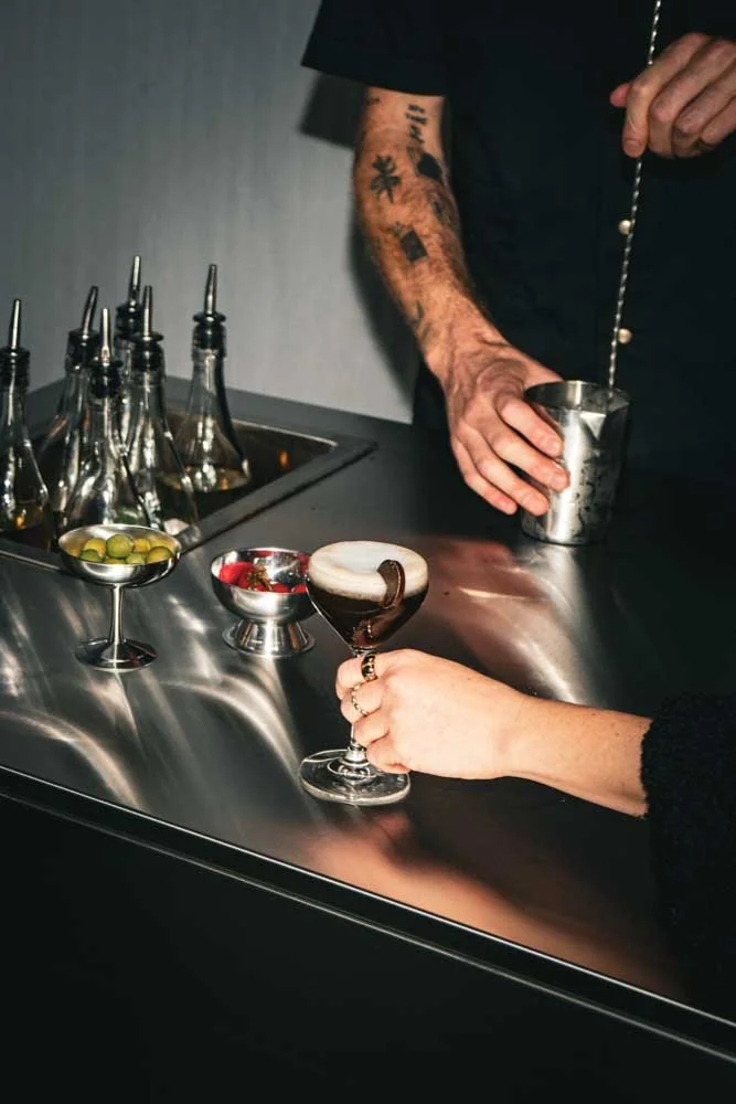 A person with tattooed arms mixing a cocktail behind a bar, while another person holds a cocktail glass with a dark drink and garnish.