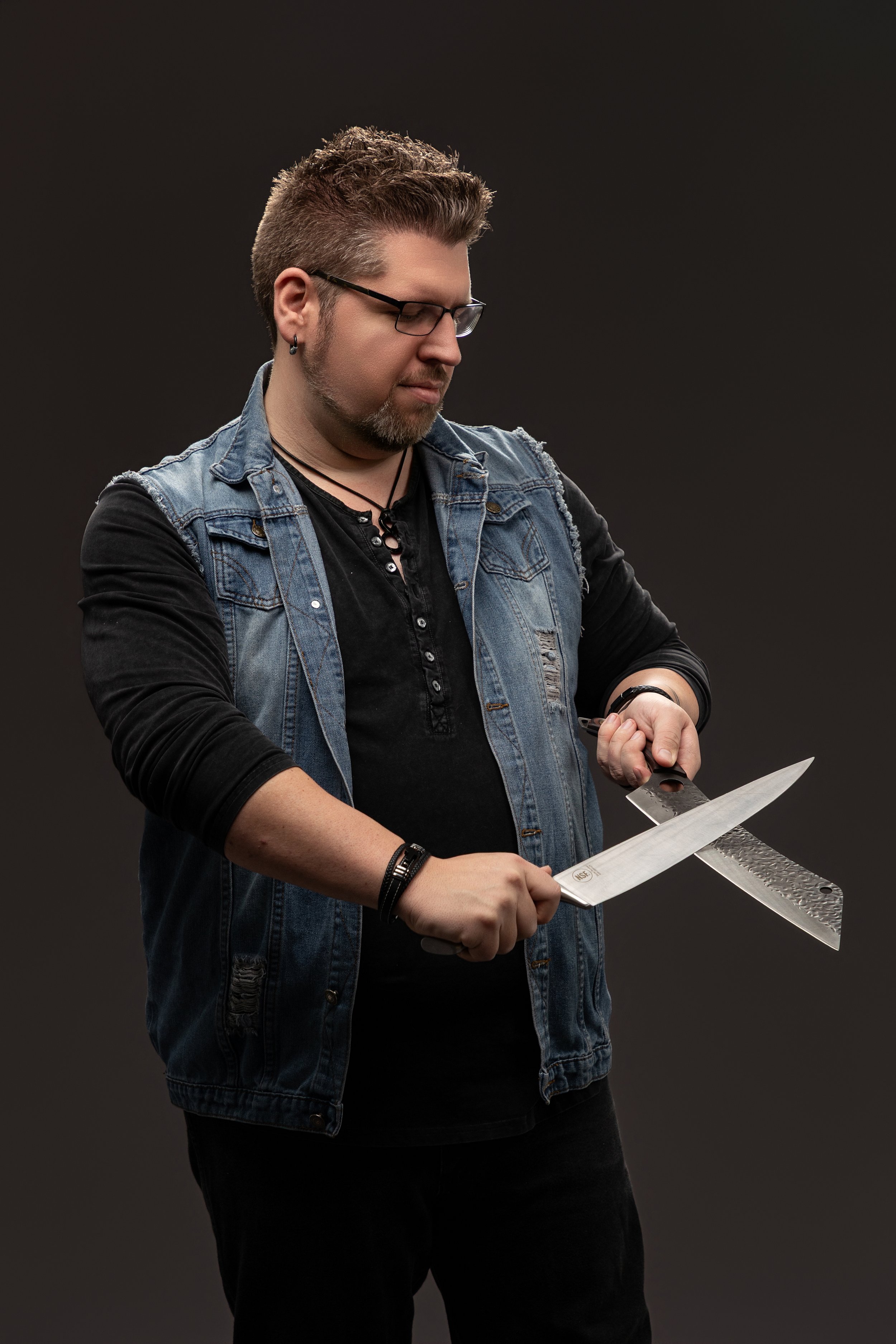 A man with glasses, a beard, and a Mohawk hairstyle holding a large kitchen knife and a cleaver against a dark background.