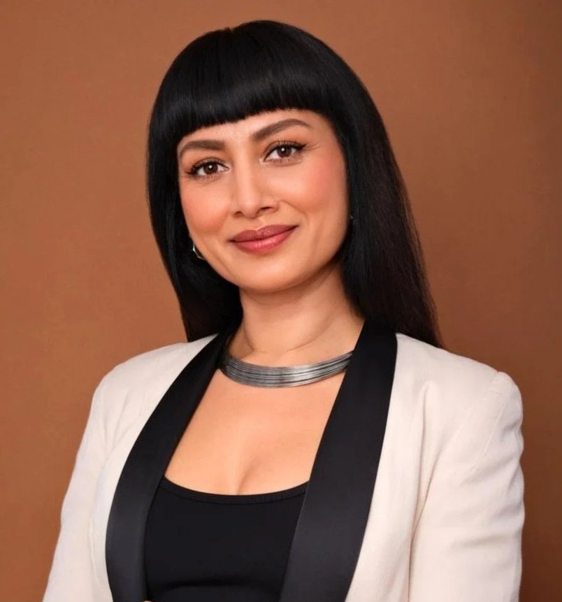 A woman with straight black hair and bangs, wearing a white blazer with black trim, a black top, and a silver necklace, smiling against a brown background.