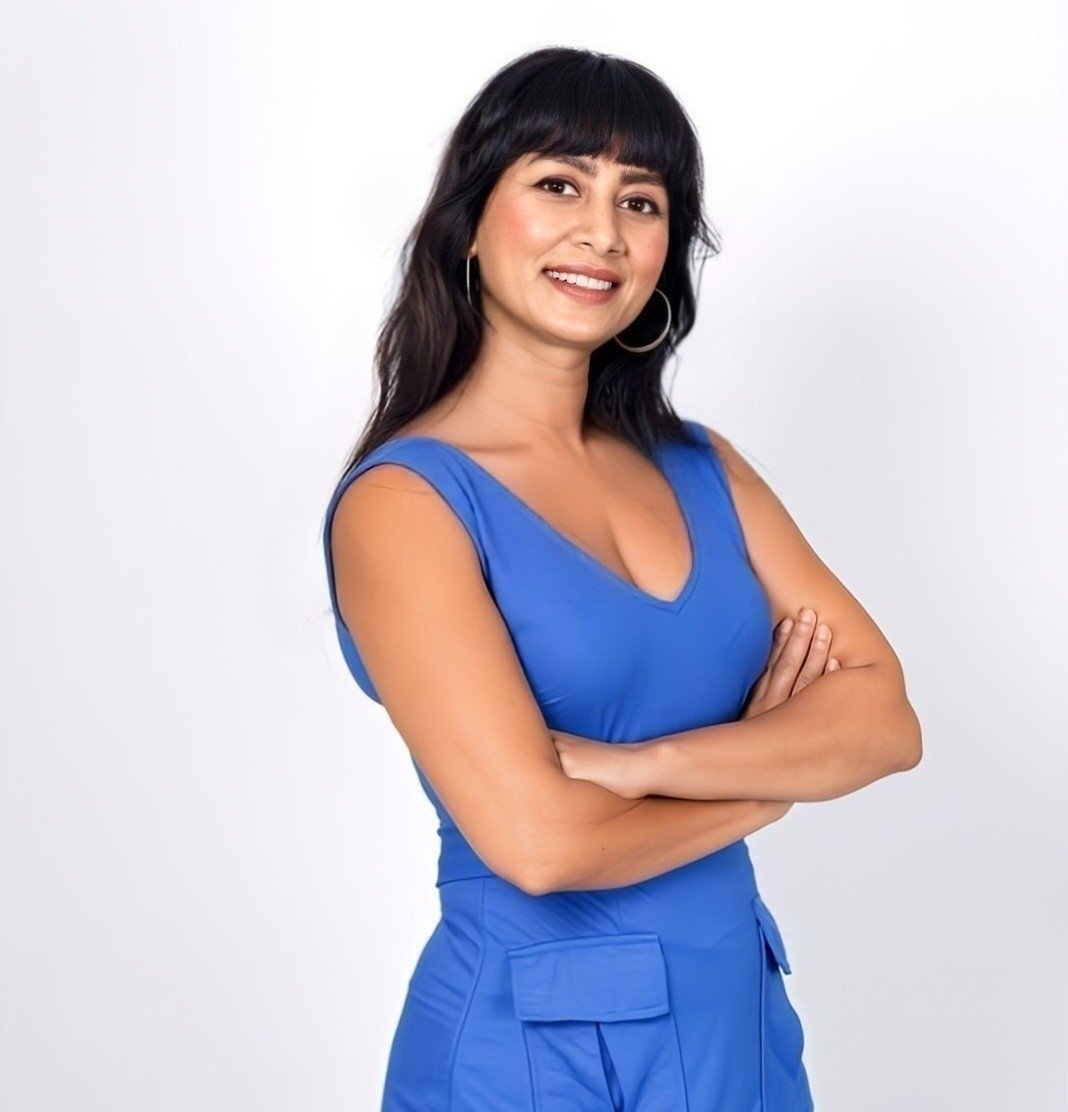 Woman with black hair and hoop earrings, smiling, wearing a blue sleeveless top and matching blue pants with pockets, standing against a white background.