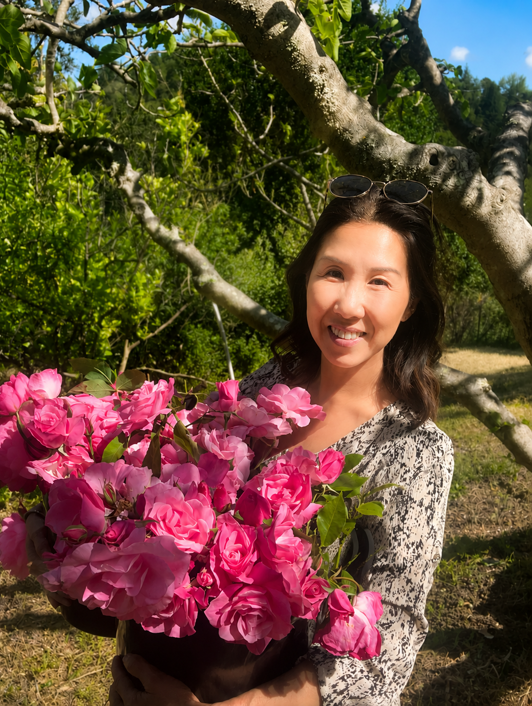 A woman smiling and holding a large pot of pink flowers outdoors, with green trees and blue sky in the background.