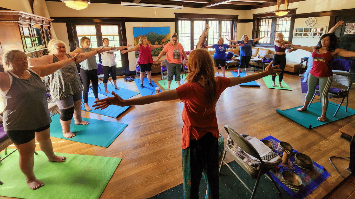 A group of women participating in a yoga or meditation class in a cozy, well-lit room with large windows, wooden floors, and traditional decor. They are standing on yoga mats with their arms extended outward, guided by an instructor in the foreground.