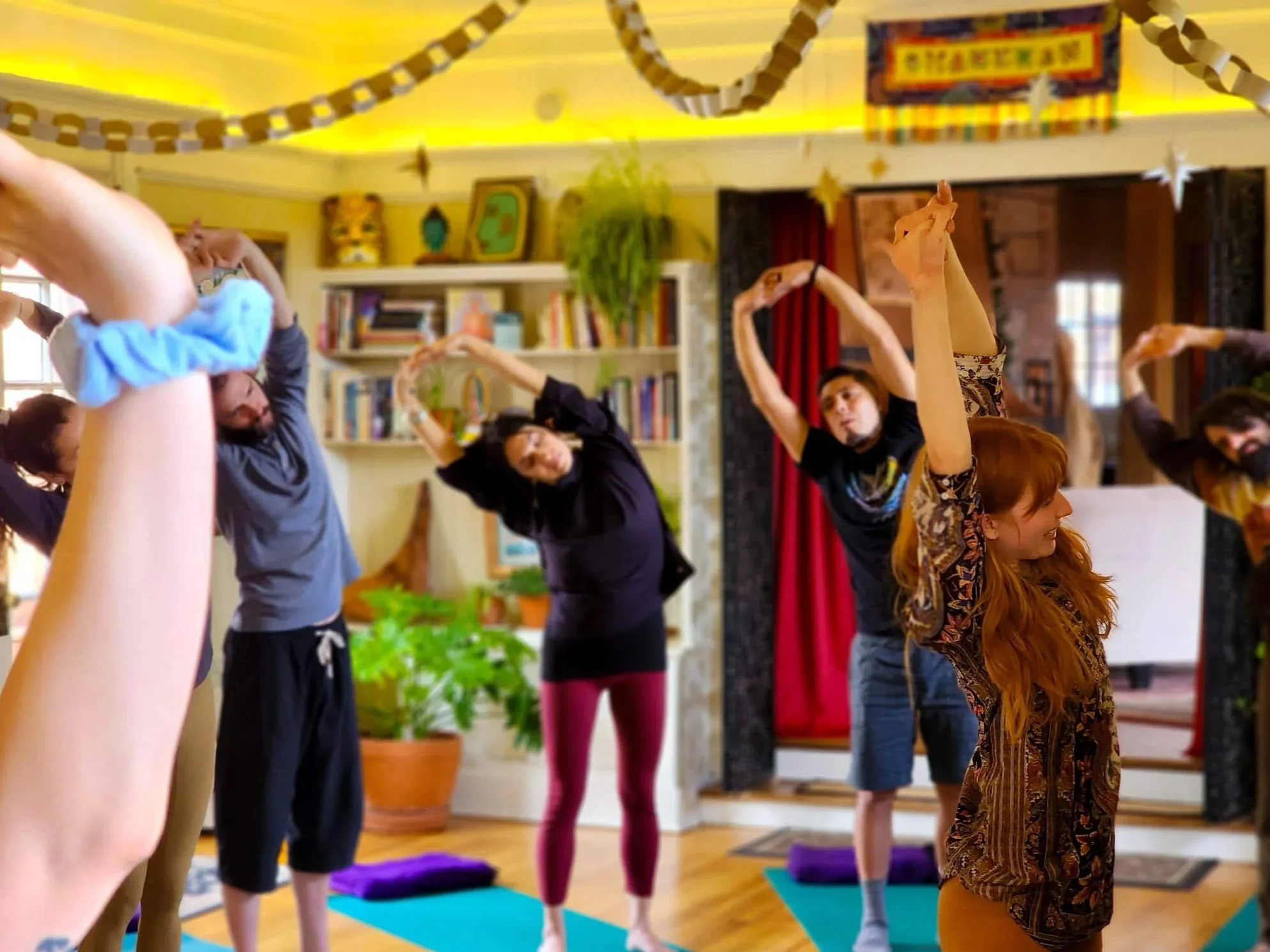 People practicing yoga in a cozy indoor space with colorful decorations, bookshelves, and a red curtain.