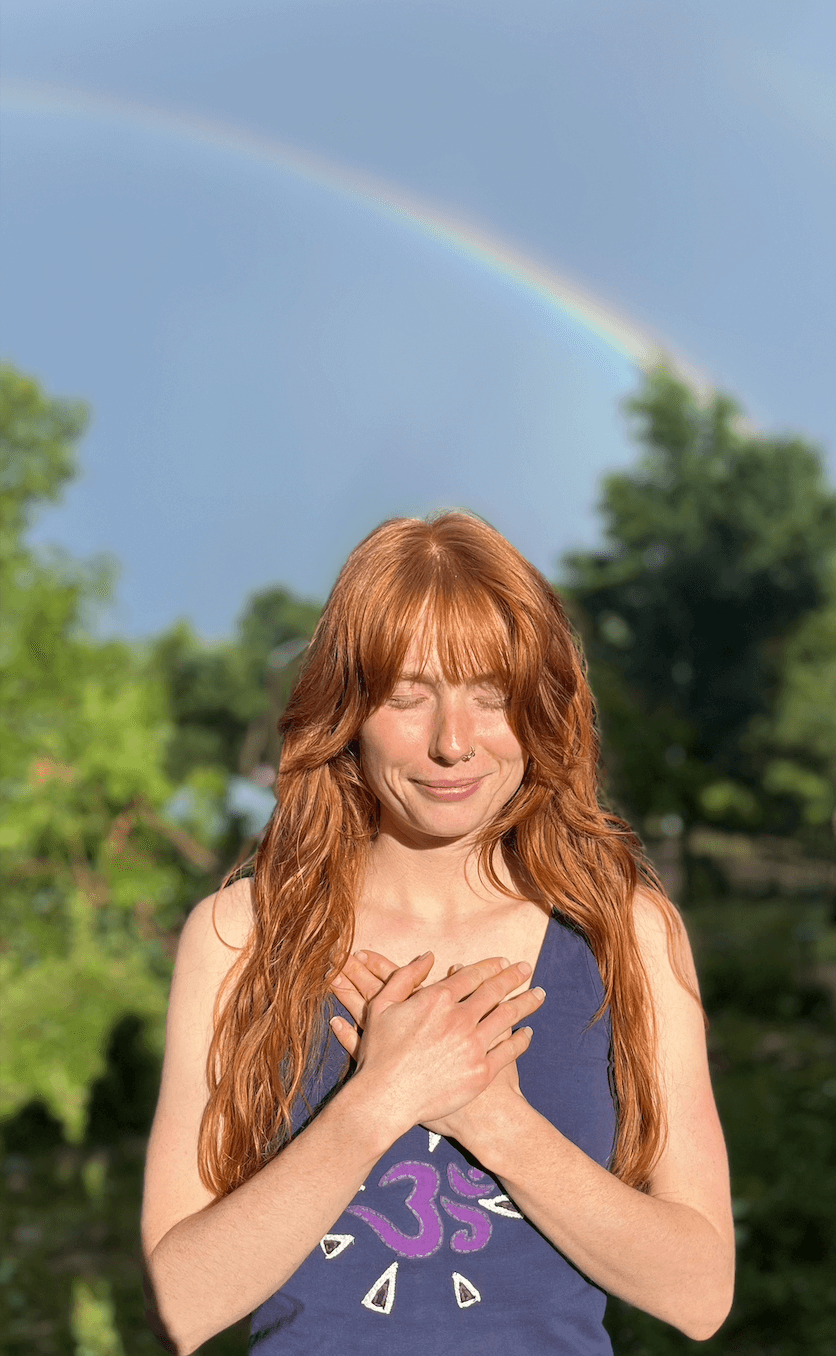 Katharine Grace Standing under a rainbow with head bowed and hands at heart.