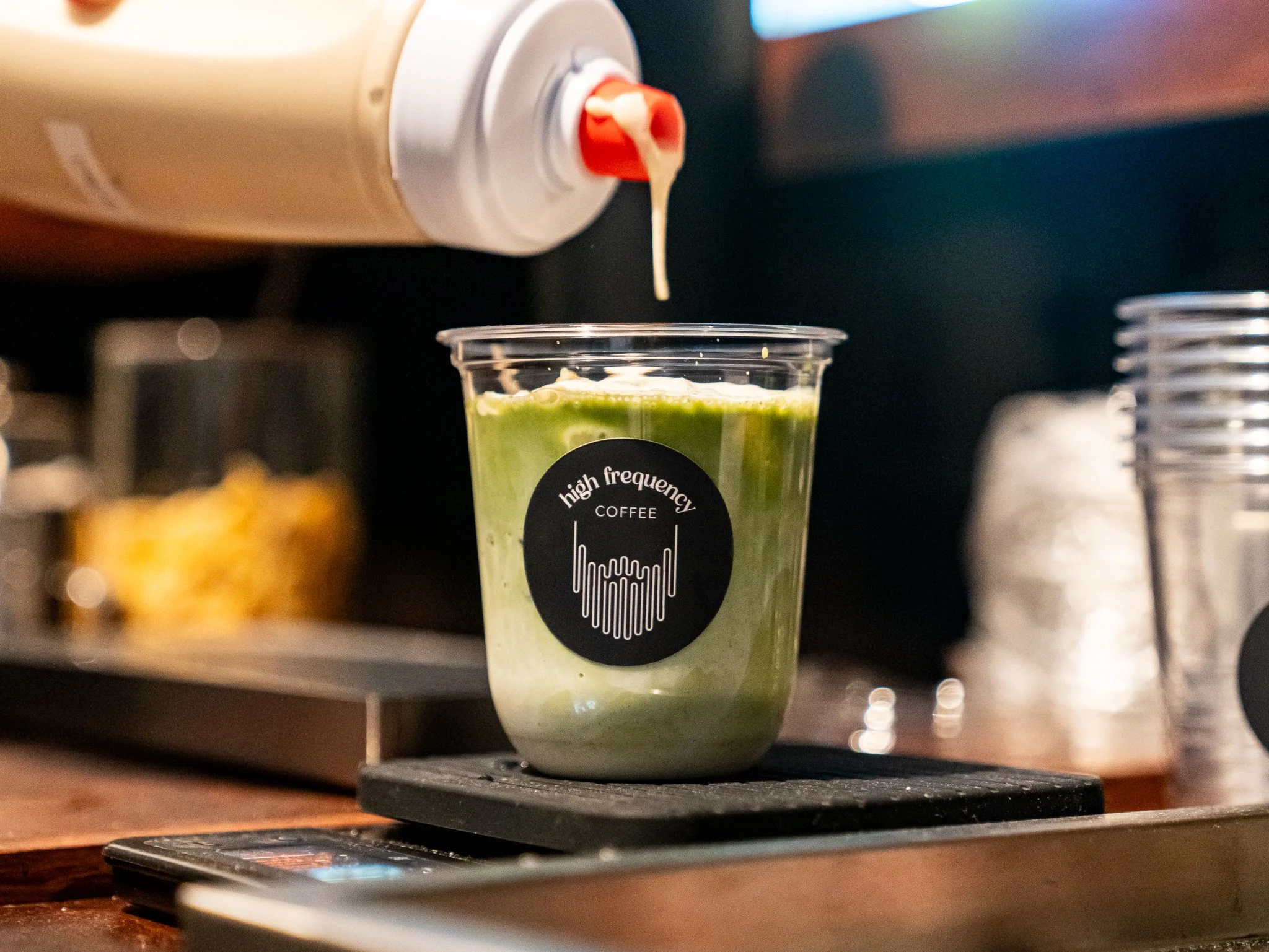 A coffee cup labeled 'high frequency' with a green beverage, possibly matcha or a smoothie, being dispensed from a white machine into the cup, with blurred background showing containers and a digital screen.