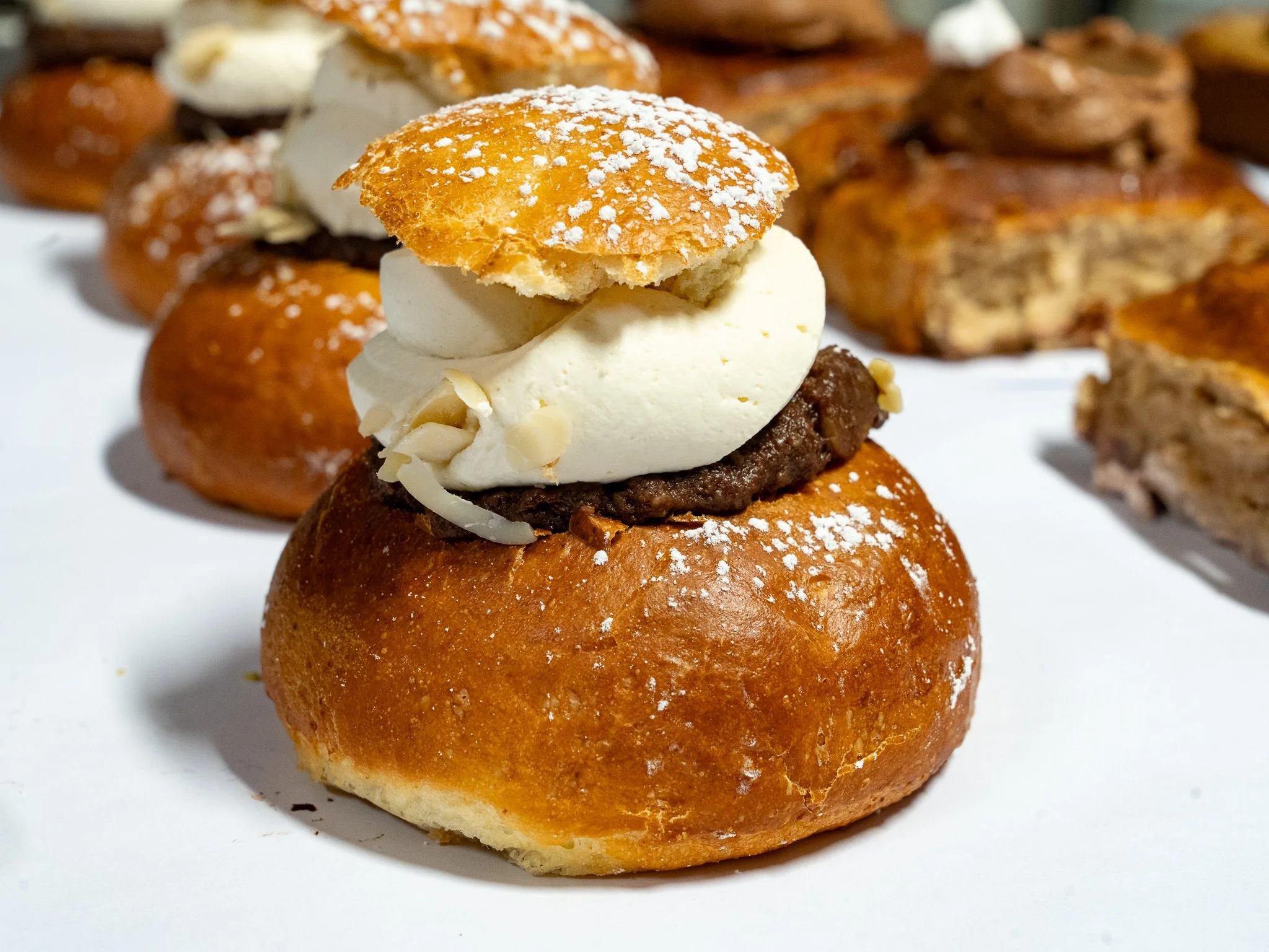 A close-up of a pastry topped with whipped cream, a dollop of chocolate filling, sliced almonds, and a small cookie, with more pastries in the background.