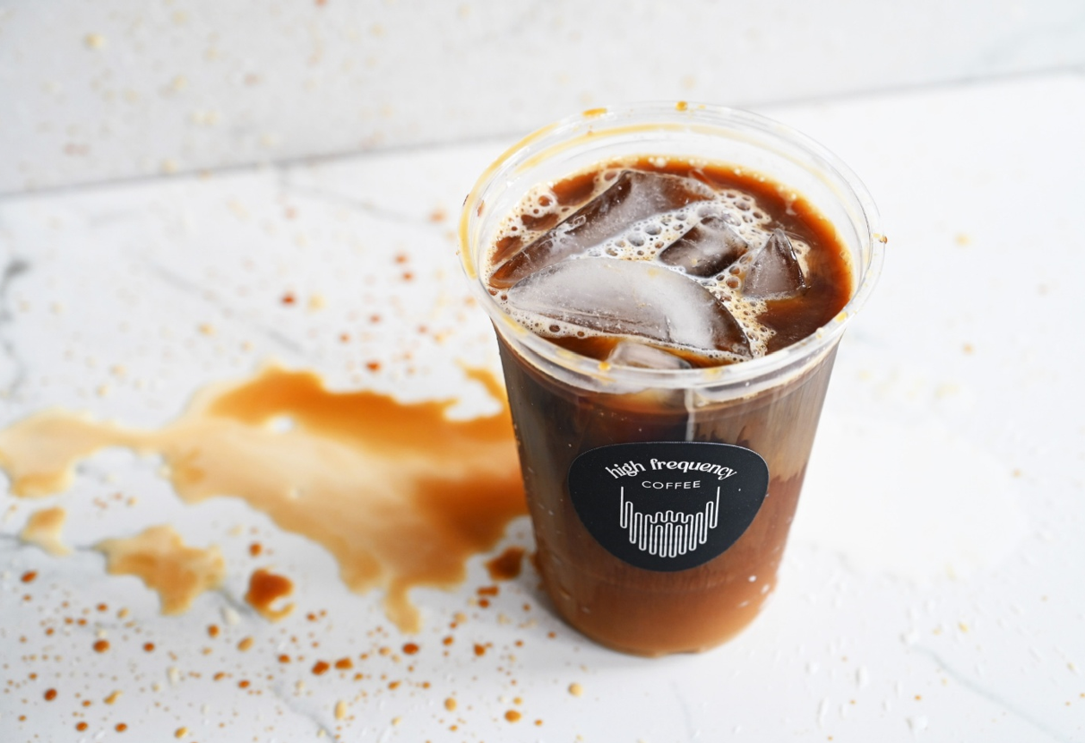 A clear plastic cup of iced coffee with a black label that says 'high frequency coffee', placed on a white surface with spilled coffee and ice cubes nearby.