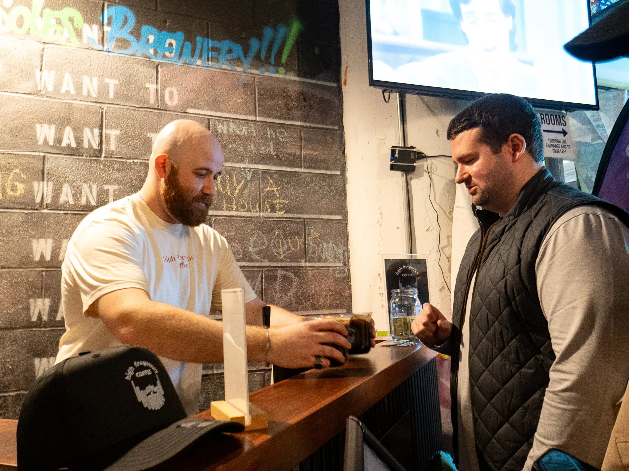 A bartender handing a drink to a customer at a bar counter in a casual setting.