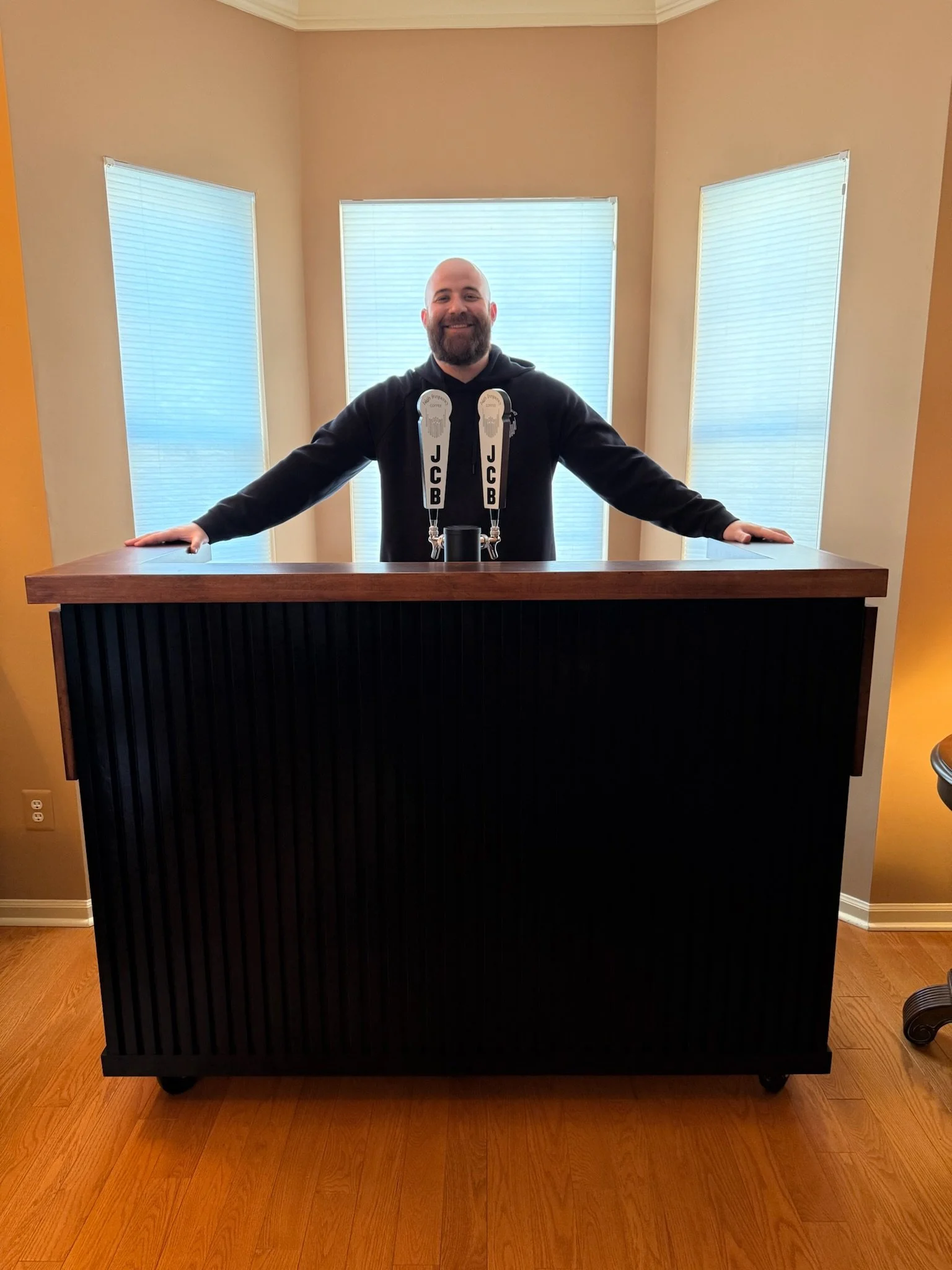 A man is standing behind a cold brew coffee tap cart. The coffee cart is elegantly design with dark wood and black paneling. He is ready to serve cold brew coffee.