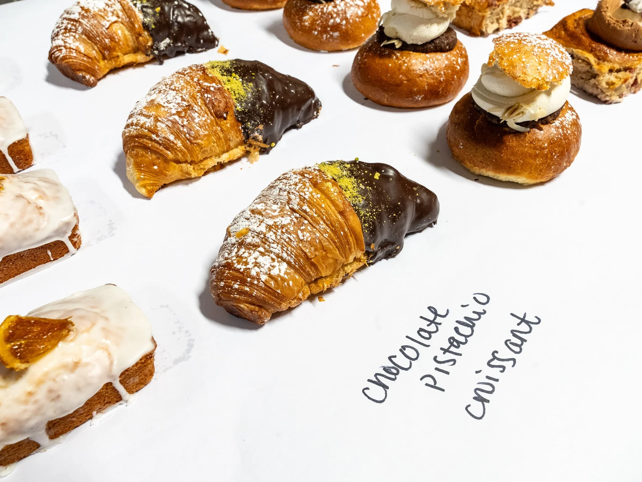 An assortment of baked goods including croissants with chocolate and pistachio, small cream-filled pastries with toppings, and a handwritten label 'Chocolate Pistachio Croissant' on a white surface.