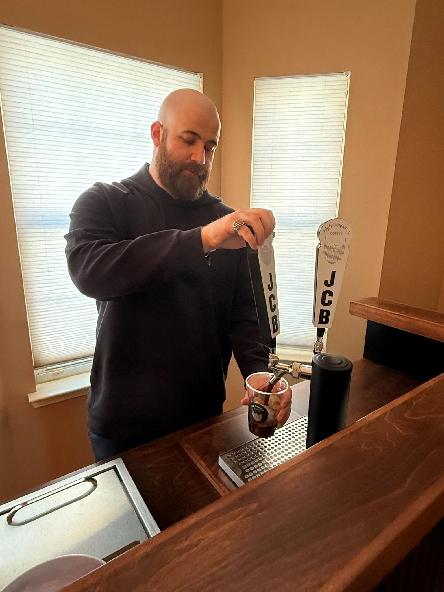 Man with beard pouring soda from a tap labeled 'JCB' into a clear plastic cup.