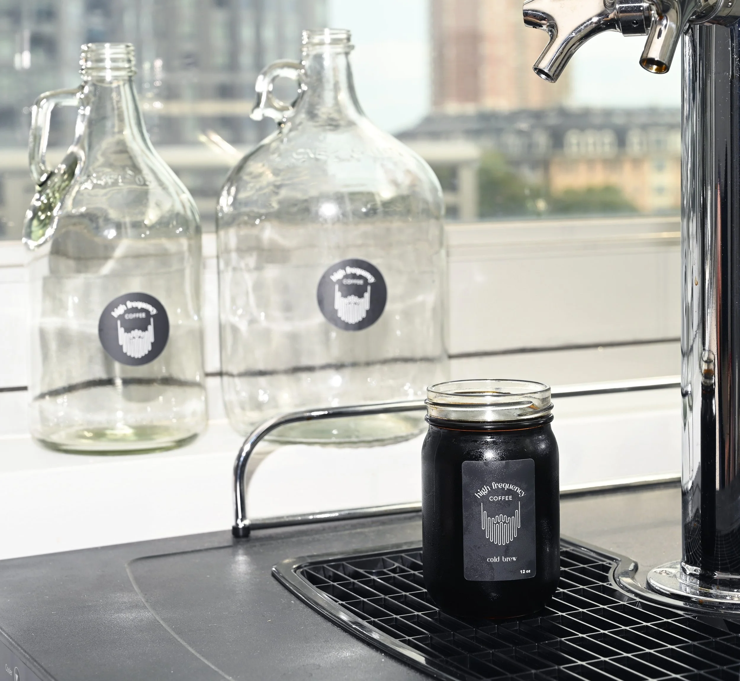 Cold brew coffee in a jar, poured from a keg tap. pictured in front of new york city. half gallon and full gallon growlers on display behind the kegerator. 