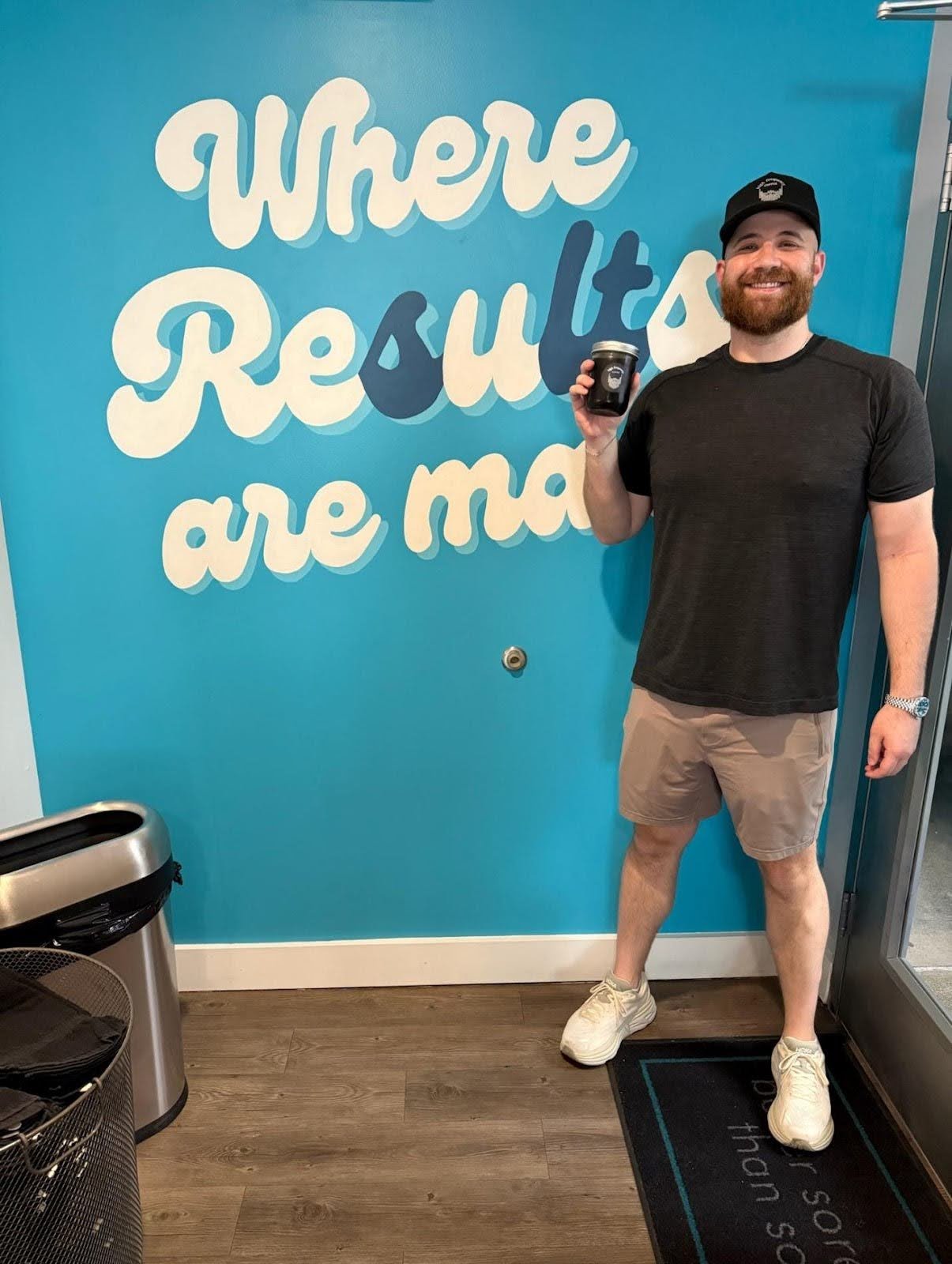 A man standing indoors in front of a turquoise wall with large white and blue text that reads 'Where Results are made.' He is smiling, wearing a black t-shirt, beige shorts, white sneakers, a watch, and a black cap. He is holding a small black cup in