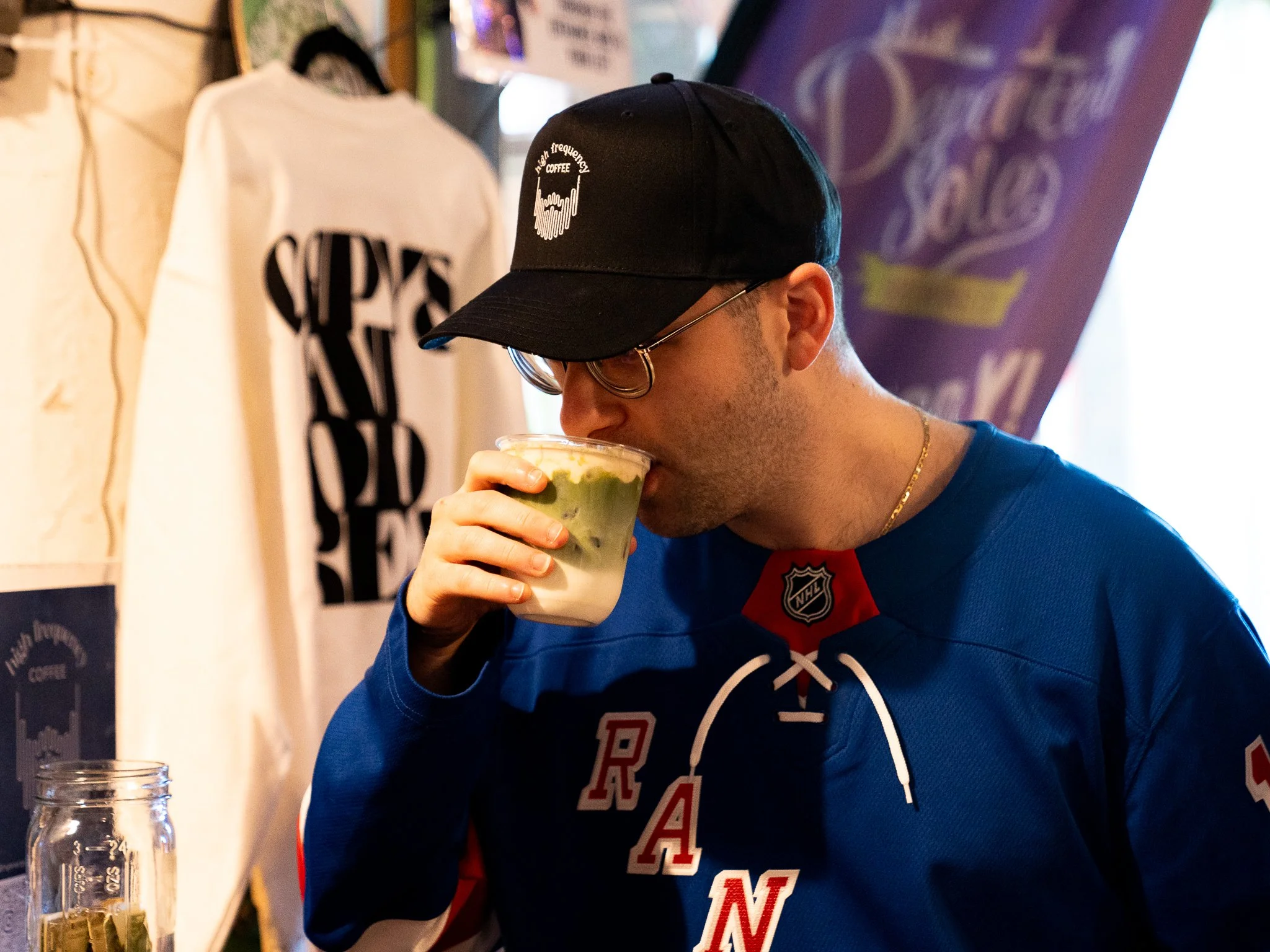 A man wearing glasses, a black cap, and a hockey jersey drinking a match with a banana cream cold foam on top.