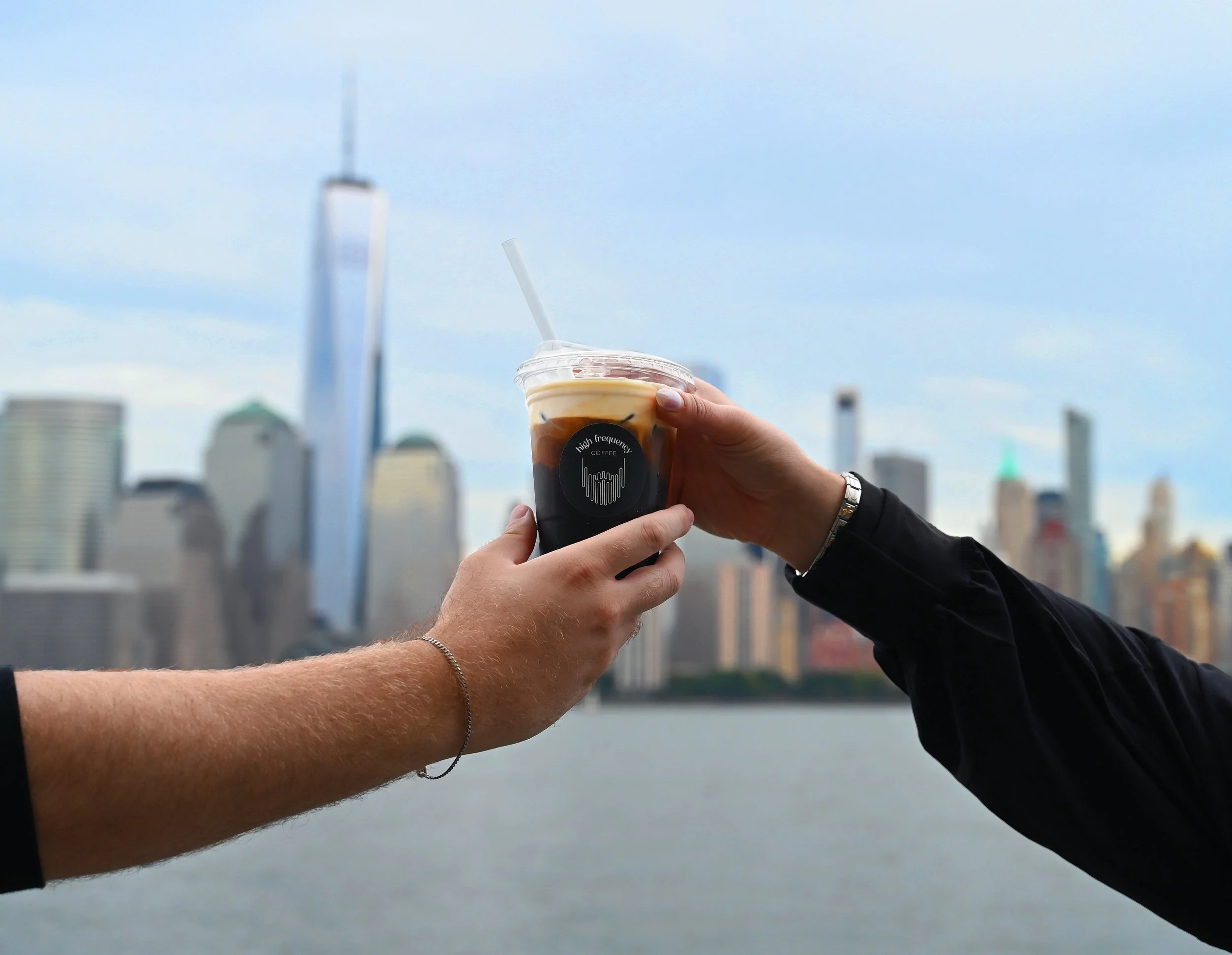 Two people are holding a cold coffee drink in a plastic cup with a straw, with a city skyline in the background across a body of water.