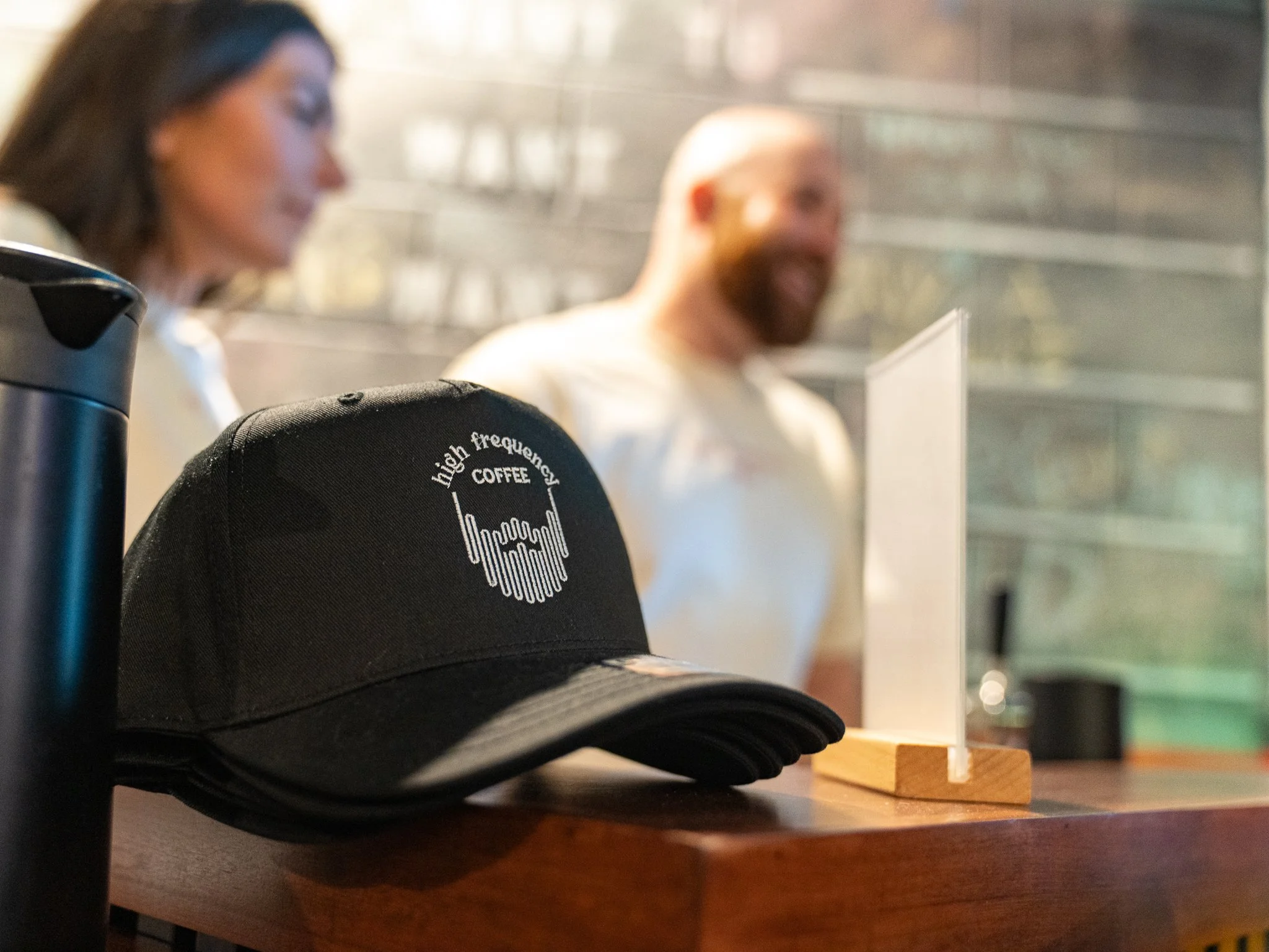 Close-up of a black baseball cap with 'high frequency coffee' logo on a wooden surface at a coffee shop, with two blurred customers in the background, one women and one man, behind a protective plexiglass divider.
