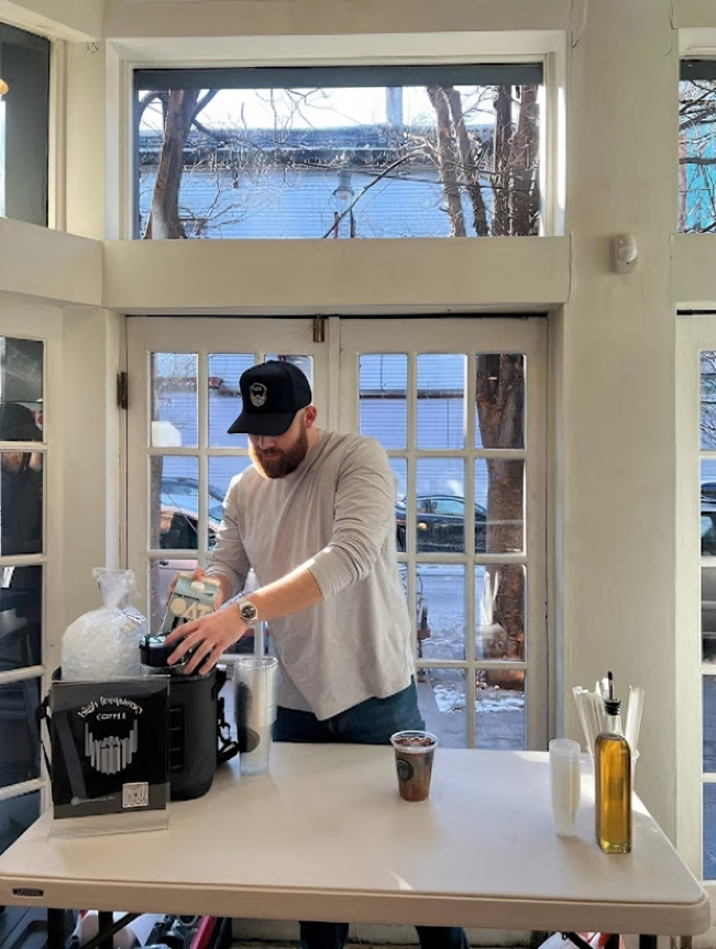 A man wearing a black cap and beige long-sleeve shirt preparing a coffee or beverage at a counter inside a cafe. Outside the window, snow and trees are visible, indicating winter weather.