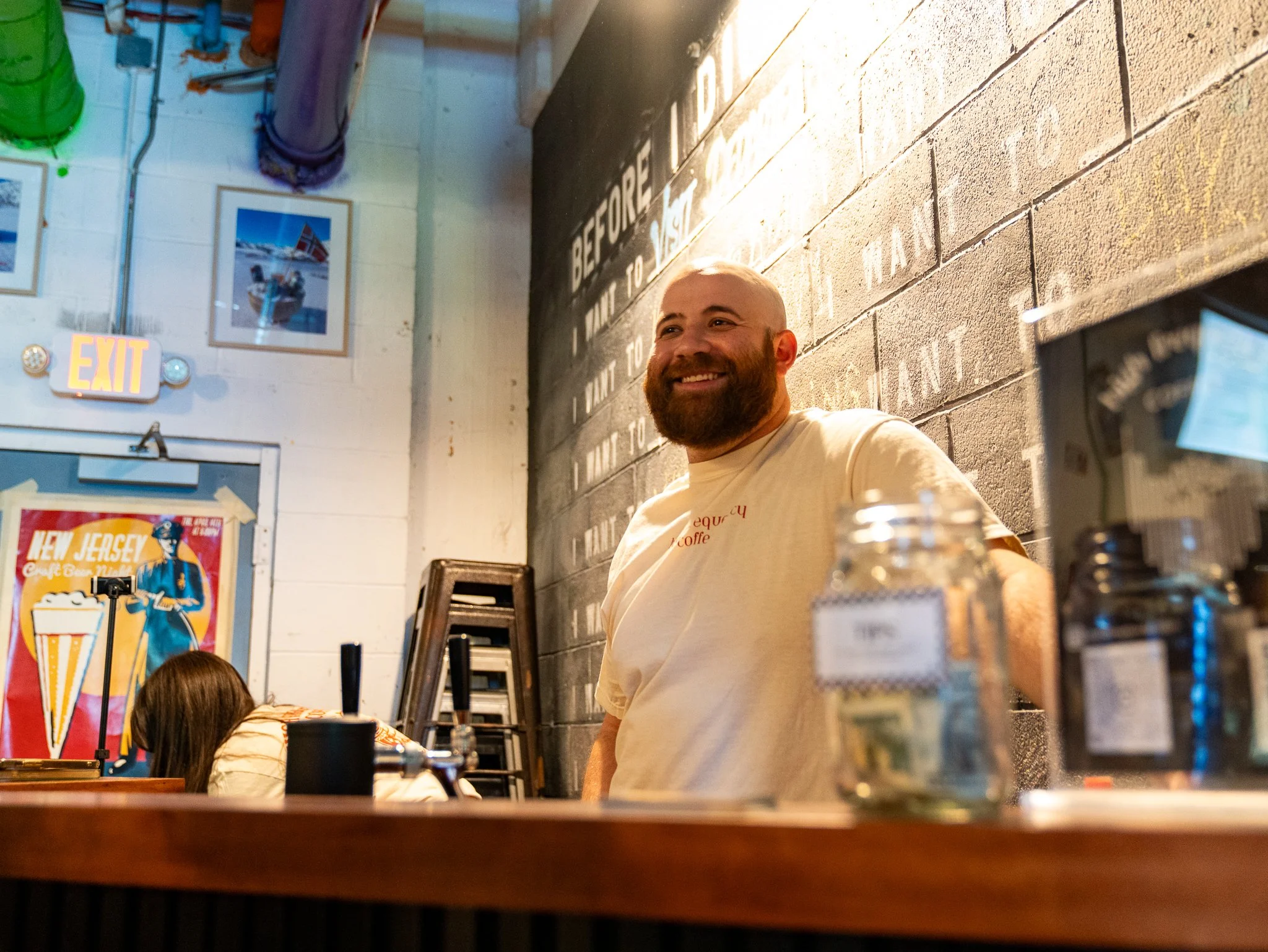 A smiling man with a beard standing behind a counter in a cafe or bar, with a woman working or cleaning in the background. The setting has colorful artwork on the wall, including a large poster of a beer, and a chalkboard with writing. There are bott