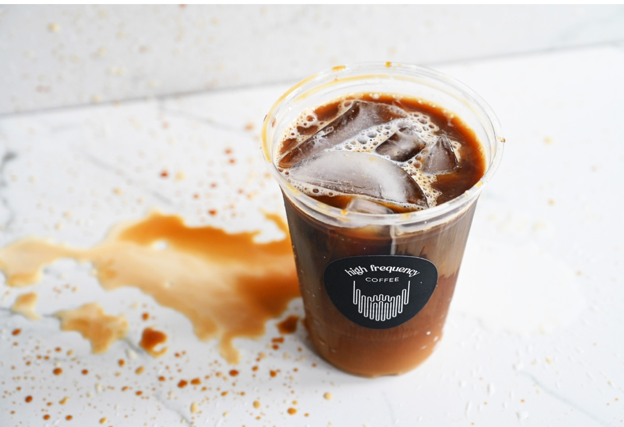 Plastic cup of iced coffee with ice cubes, spilled coffee stain on white surface, and a gray background, with a logo that says 'high frequency COFFEE'.