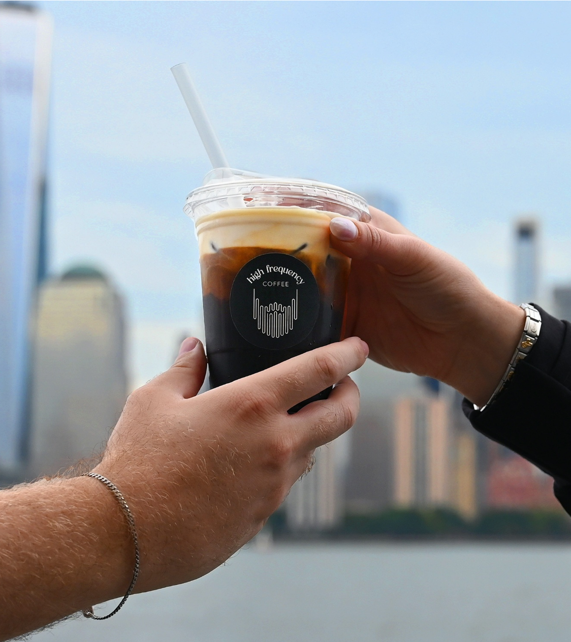 Two hands holding a plastic cup of iced coffee with a logo that reads 'high frequency COFFEE' and a city skyline in the background.