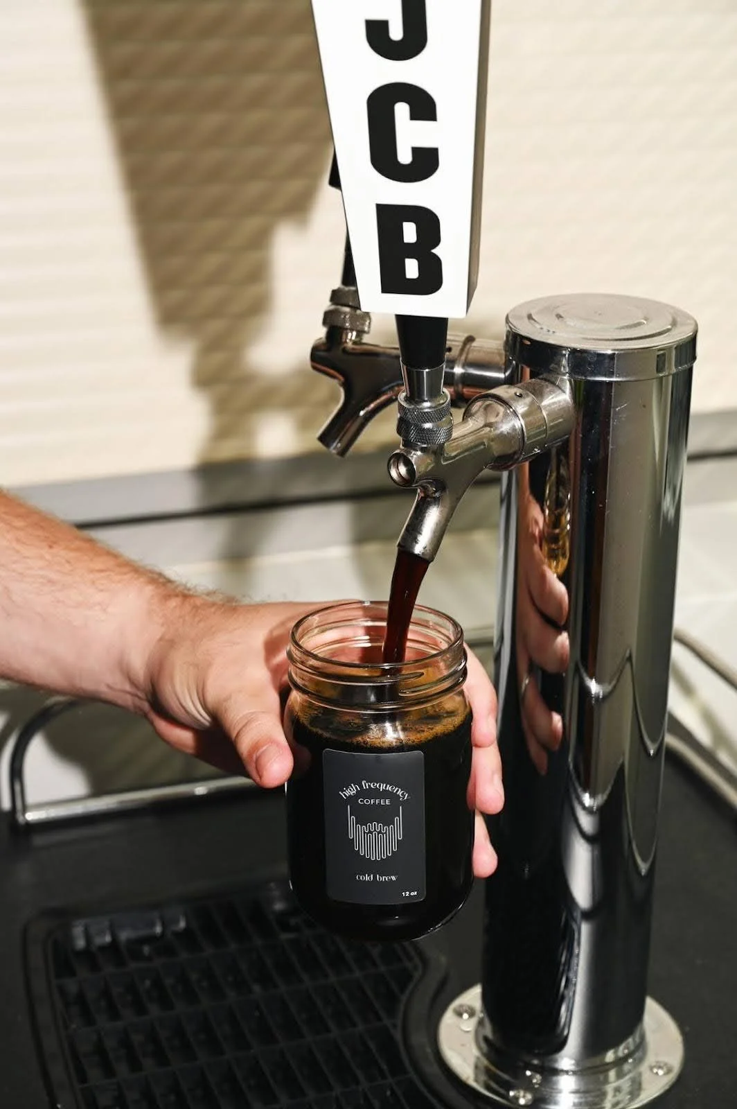 A hand filling a glass jar with cold brew coffee from a coffee tap labeled 'JCB' at a café or coffee station.