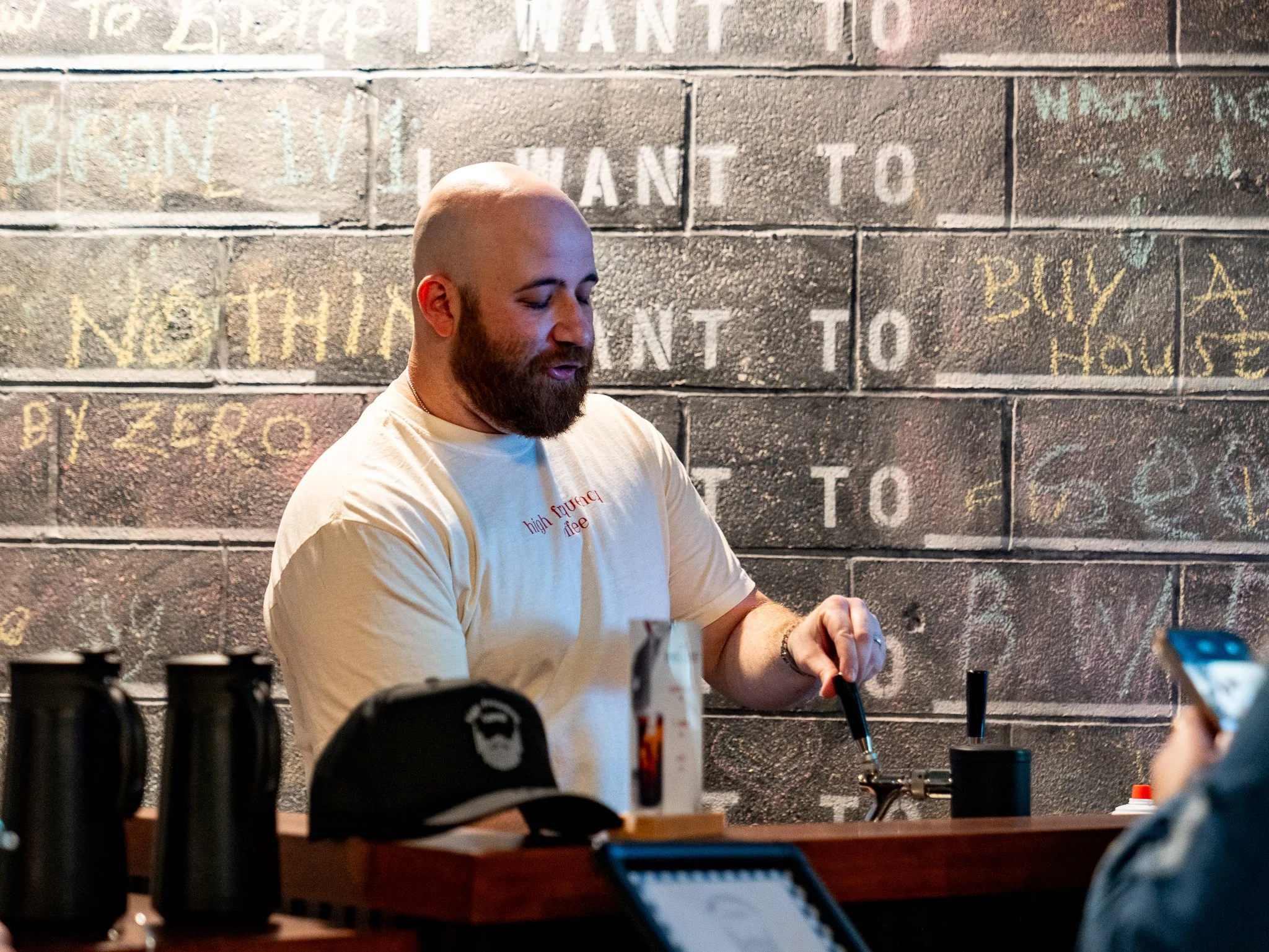 A man with a beard and shaved head making coffee at a coffee shop counter, with chalk writing on a brick wall behind him.