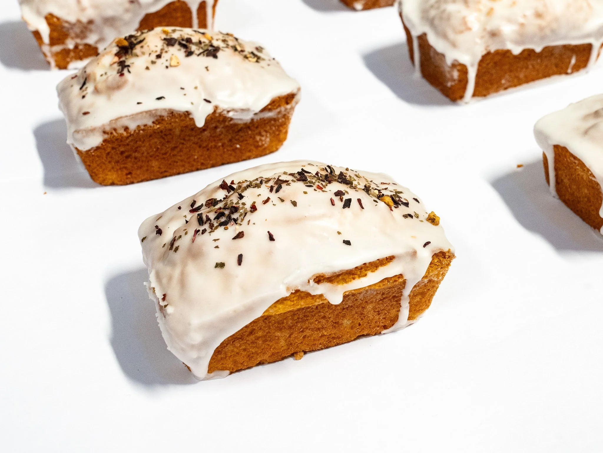 Four rectangular pieces of carrot cake with white frosting and sprinkled with chopped nuts and herbs on a white background.