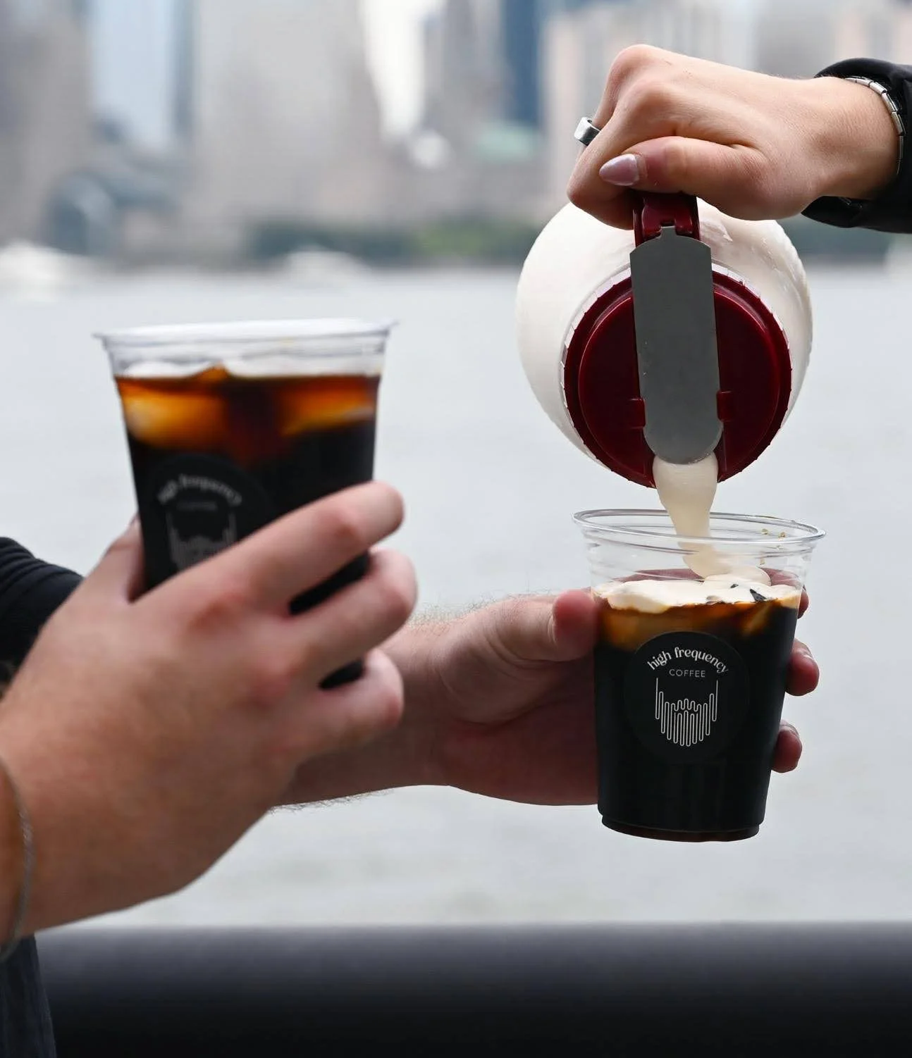 Person pouring creamer into a plastic cup of black coffee while another person holds a similar coffee cup.