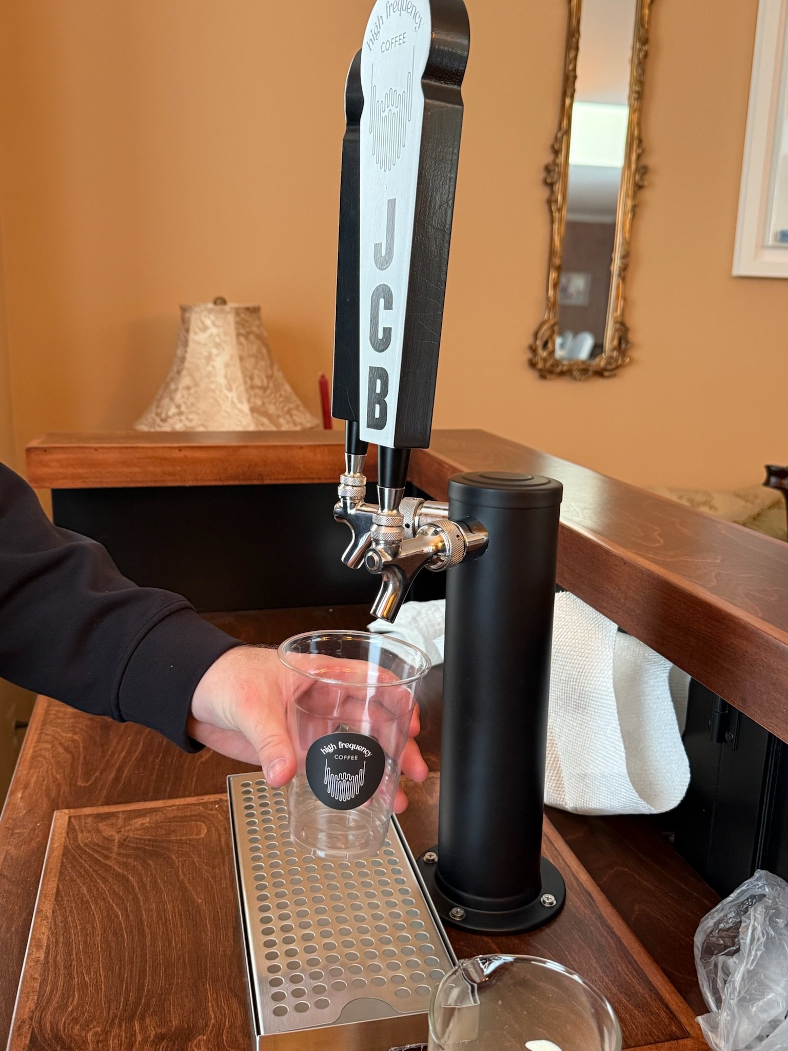A person is filling a clear plastic cup from a tap attached to a black beverage dispenser with foamed coffee labeled "High Frequency Coffee." The setting appears to be indoors in a room with beige walls, featuring a decorative mirror and a table lamp in the background.