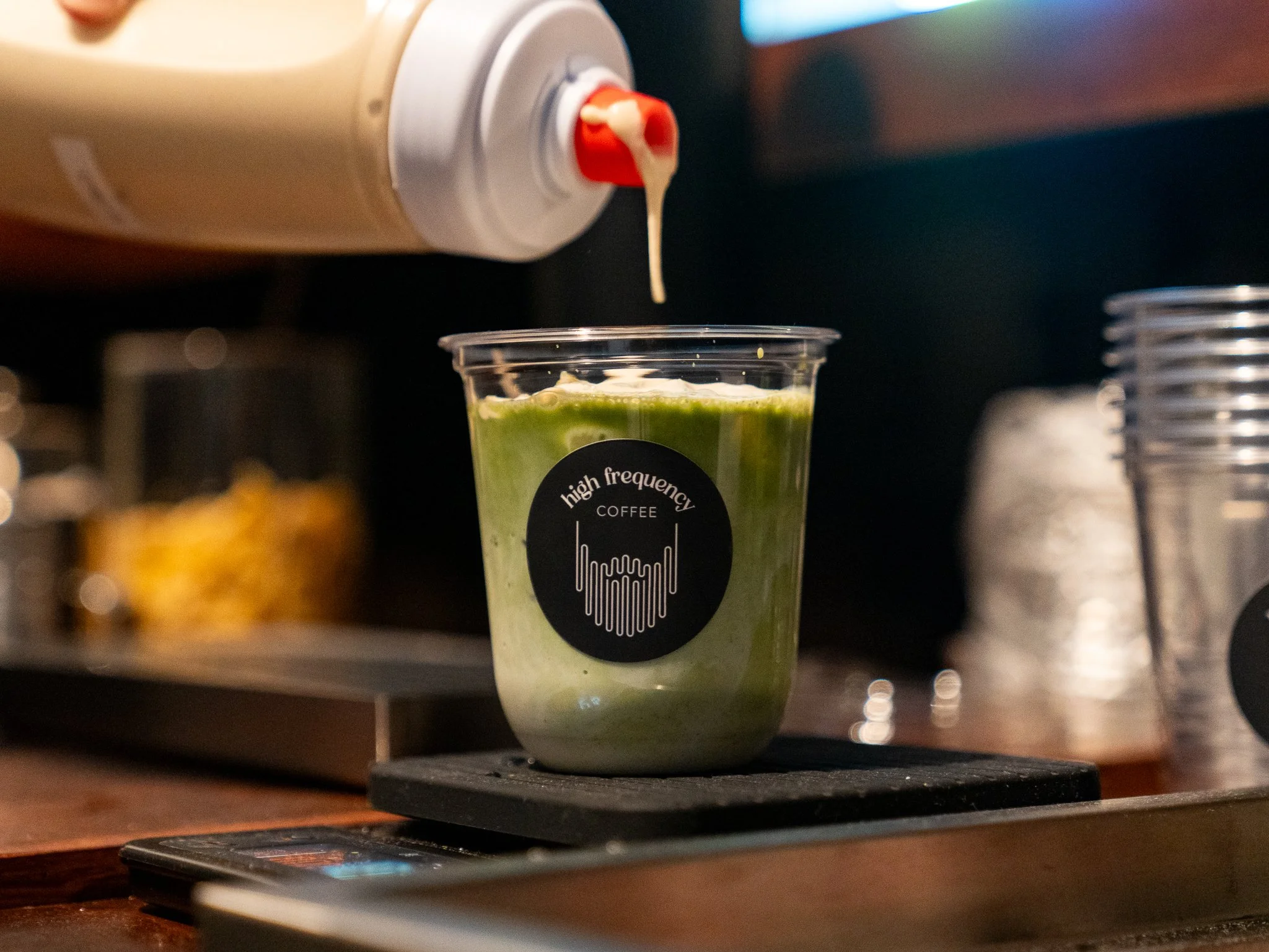 A plastic cup with a matcha, labeled 'high frequency COFFEE,' is placed on a scale with a background of coffee equipment and disposable cups.