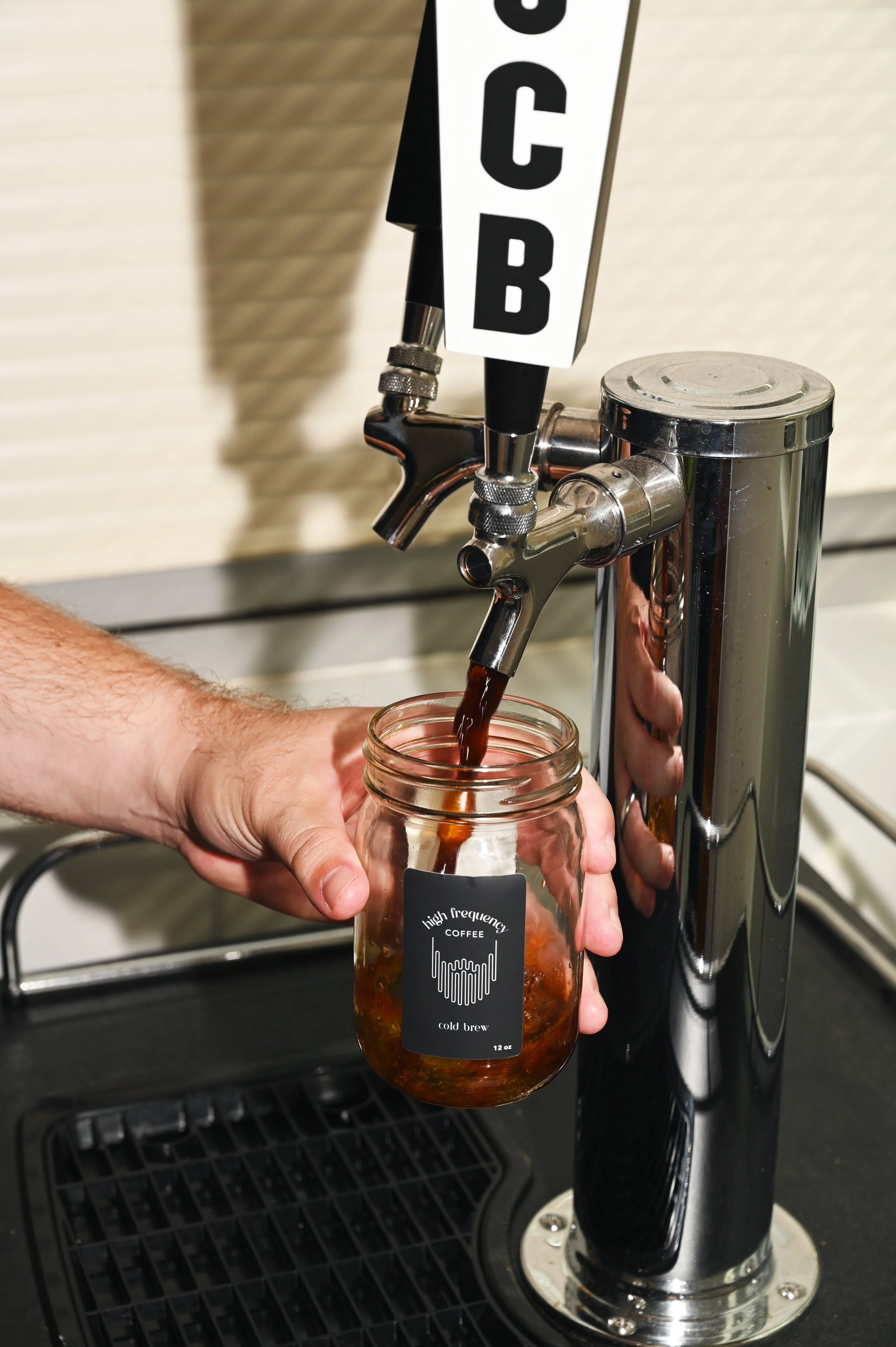 Person filling a glass jar with dark cold brew coffee from a chrome tap labeled 'COFFEE'.
