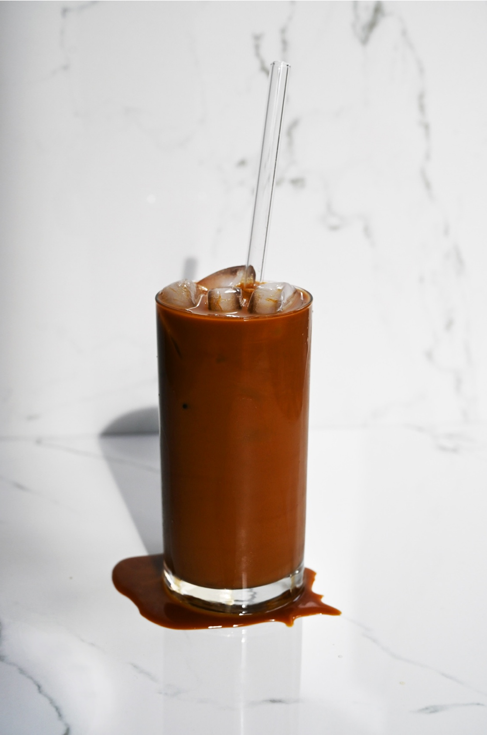 A tall glass of iced coffee with a straw on a white marble surface, with spilled coffee at the base of the glass.